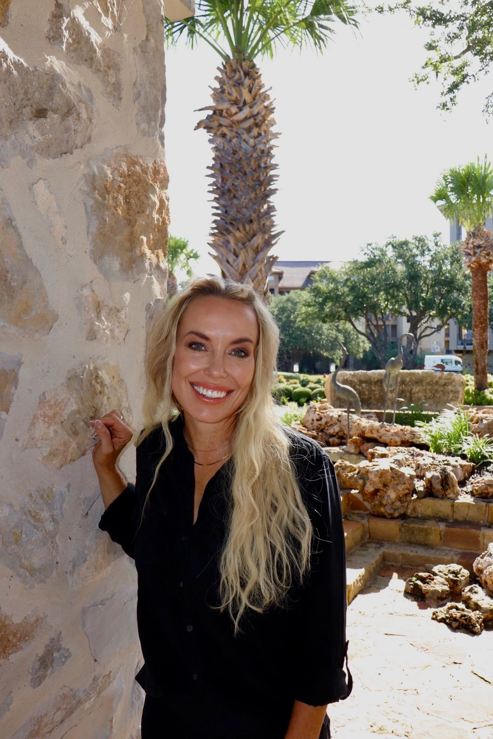 A smiling woman with long blonde hair standing outdoors near a stone wall, with palm trees and decorative flamingo statues in the background.