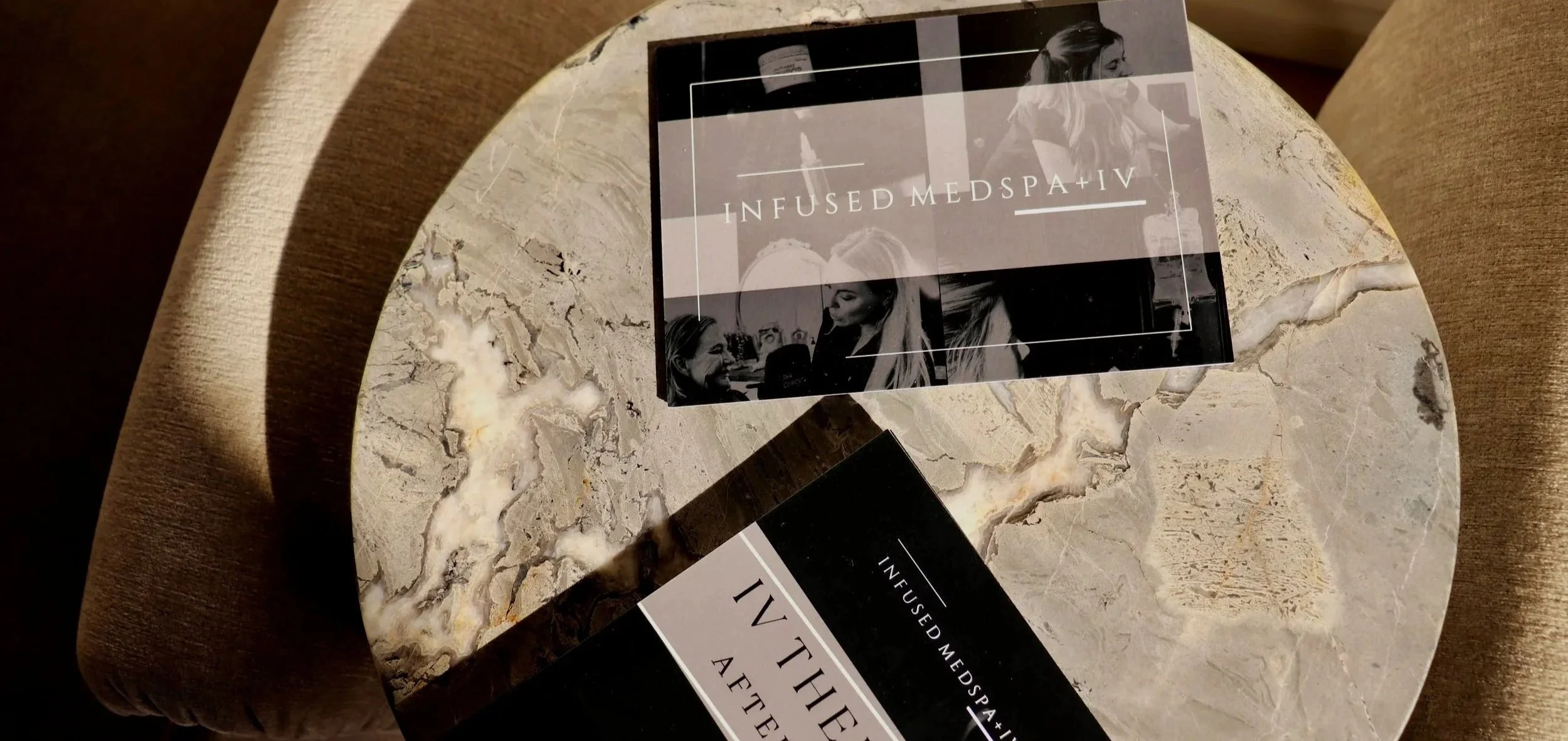 Two black and white pamphlets titled "Infused Medspa+IV" resting on a marble table, with a woman's face visible on the cover of one pamphlet.