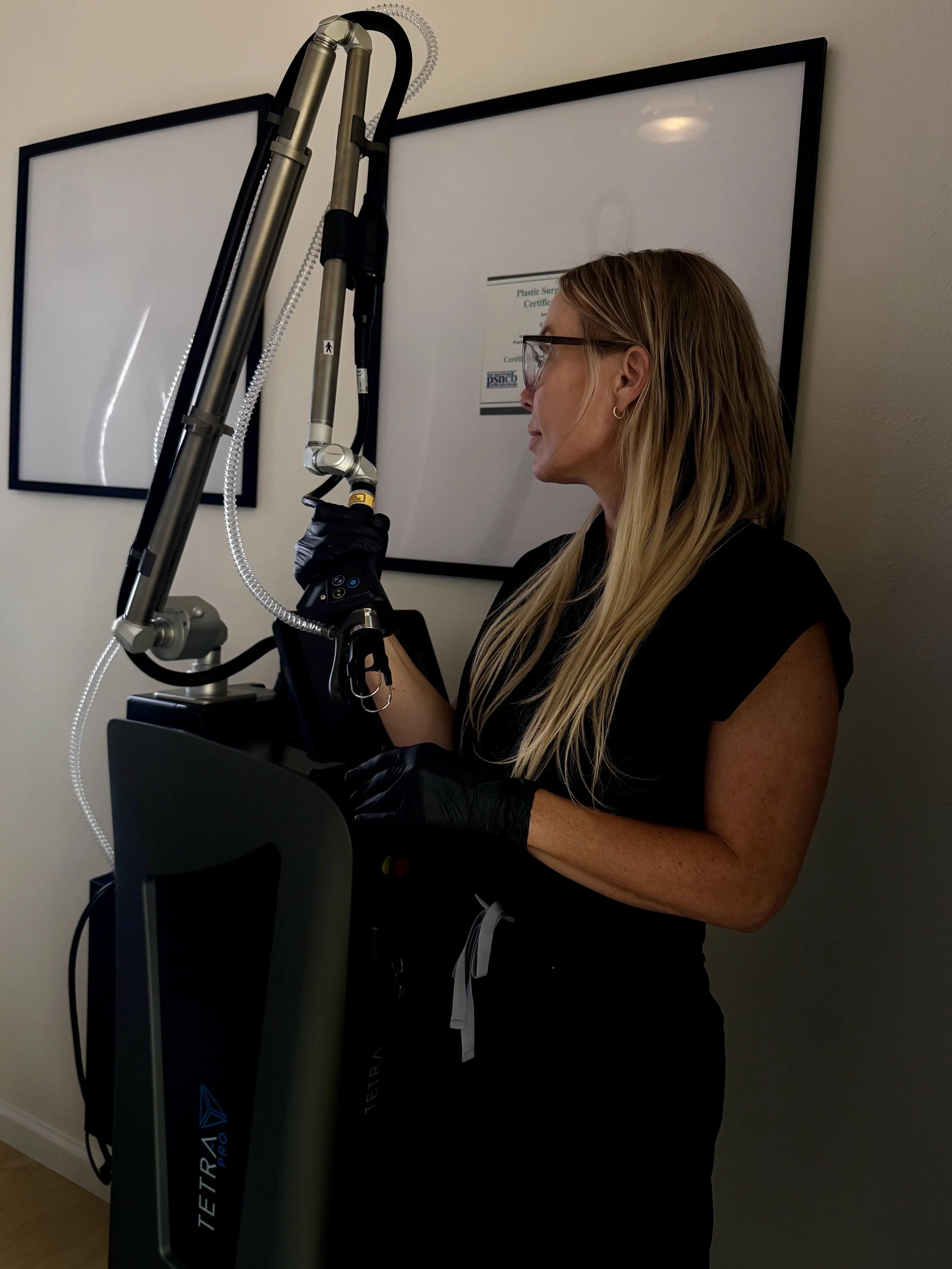 A woman with long blonde hair, wearing glasses and black gloves, stands near a Tetra laser device in a medical or professional setting.