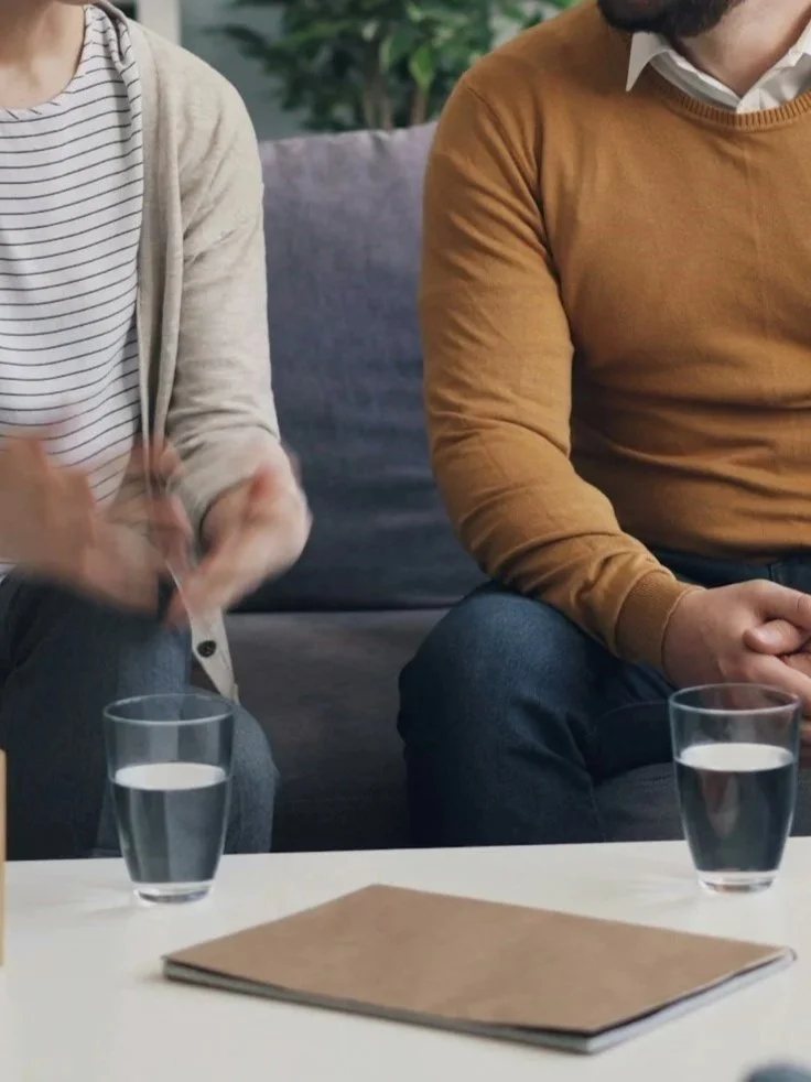 Two people sitting on a gray couch with a table in front of them, holding glasses of water. A brown folder or notebook is on the table.