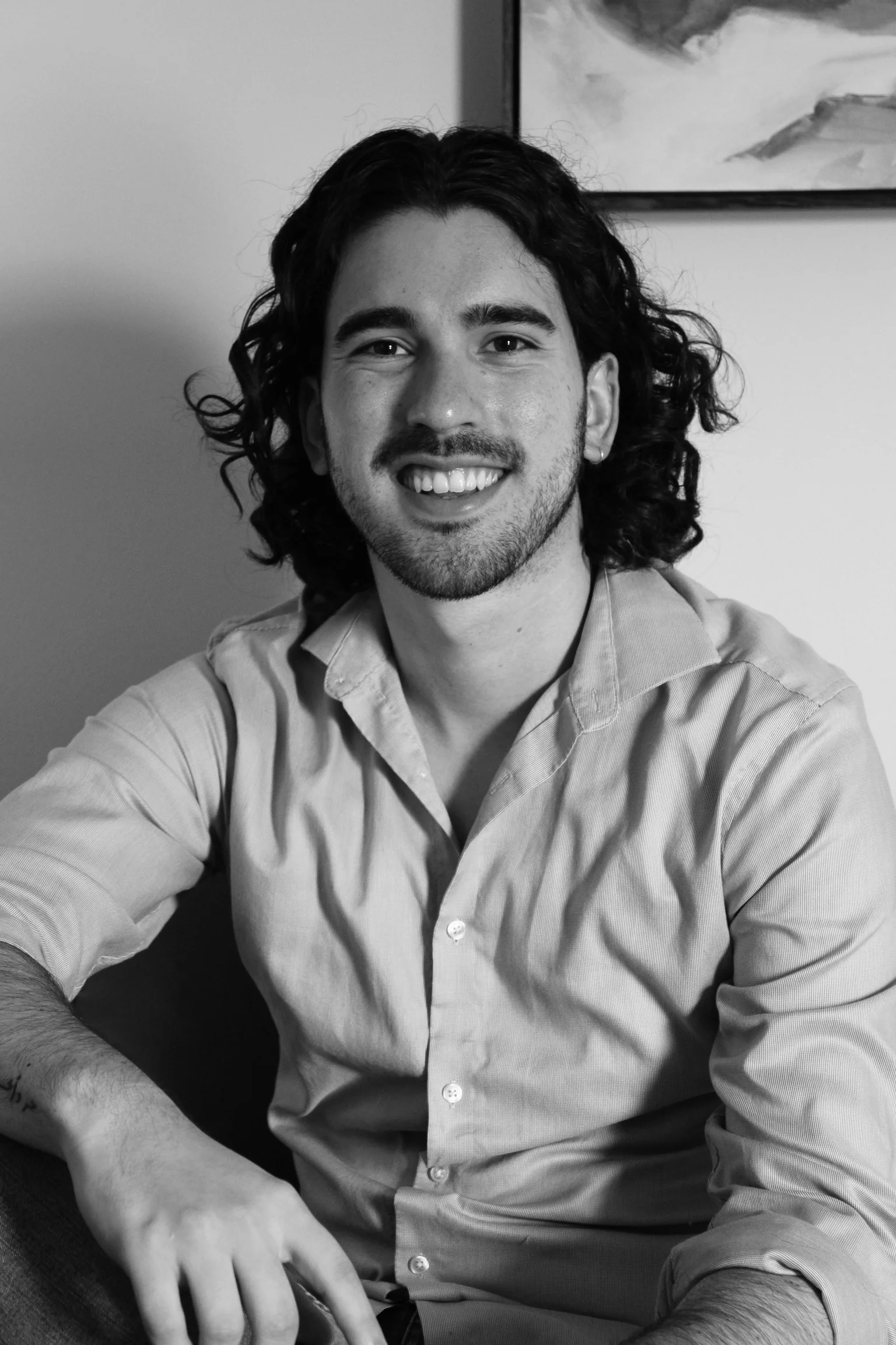 Black and white portrait of a young man with shoulder-length curly hair, smiling, wearing a light-colored button-up shirt, sitting indoors with a wall and a painting in the background.