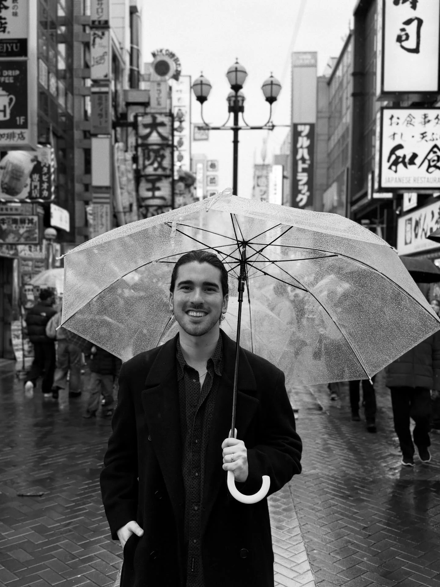 A man smiling and holding a transparent umbrella on a rainy city street with signs in foreign characters in the background.