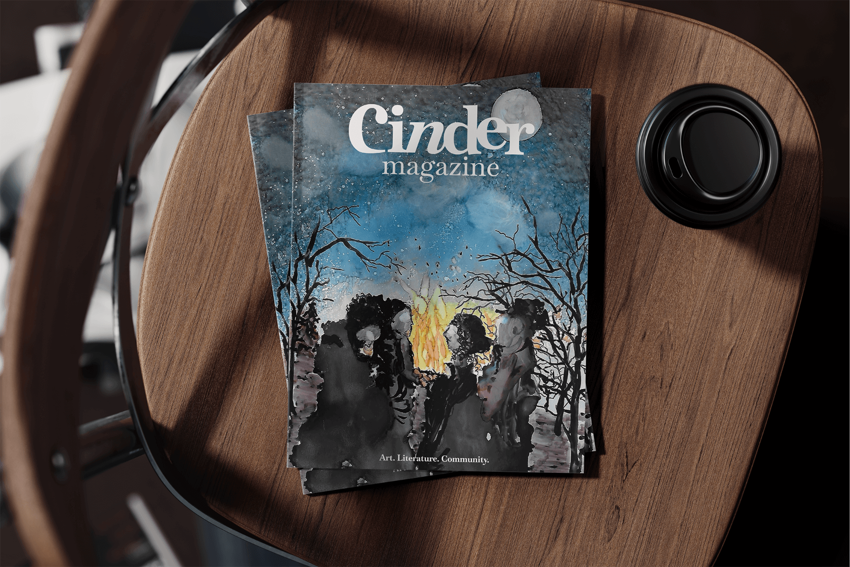 A mocked up image of 2 print issues of Cinder Magazine Issue No. 1, placed on a chair next to a coffee cup.