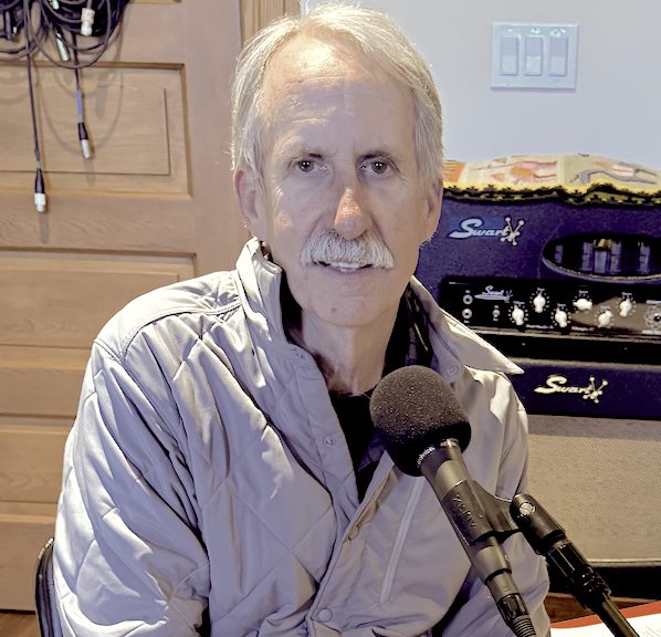 An older man with gray hair and a mustache sits in front of a microphone, wearing a light gray quilted jacket, in a room with a wooden cabinet, musical equipment, and a guitar amplifier.