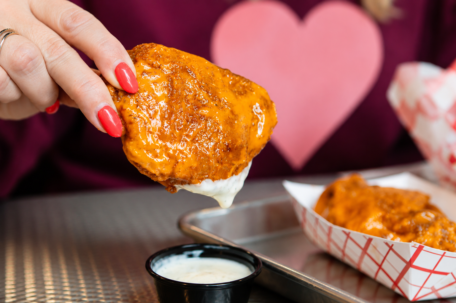 Close-up of a hand with pink painted nails holding a buffalo style sauced chicken thigh aka "Thwang", in front of a pink heart-shaped decoration and a tray with more "Thwang" chicken thighs and a small container of dipping sauce.