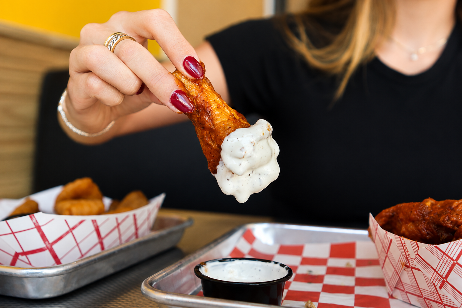 Person dipping a chicken wing into ranch dipping sauce with compensation to a tray of chicken wings in a basket.