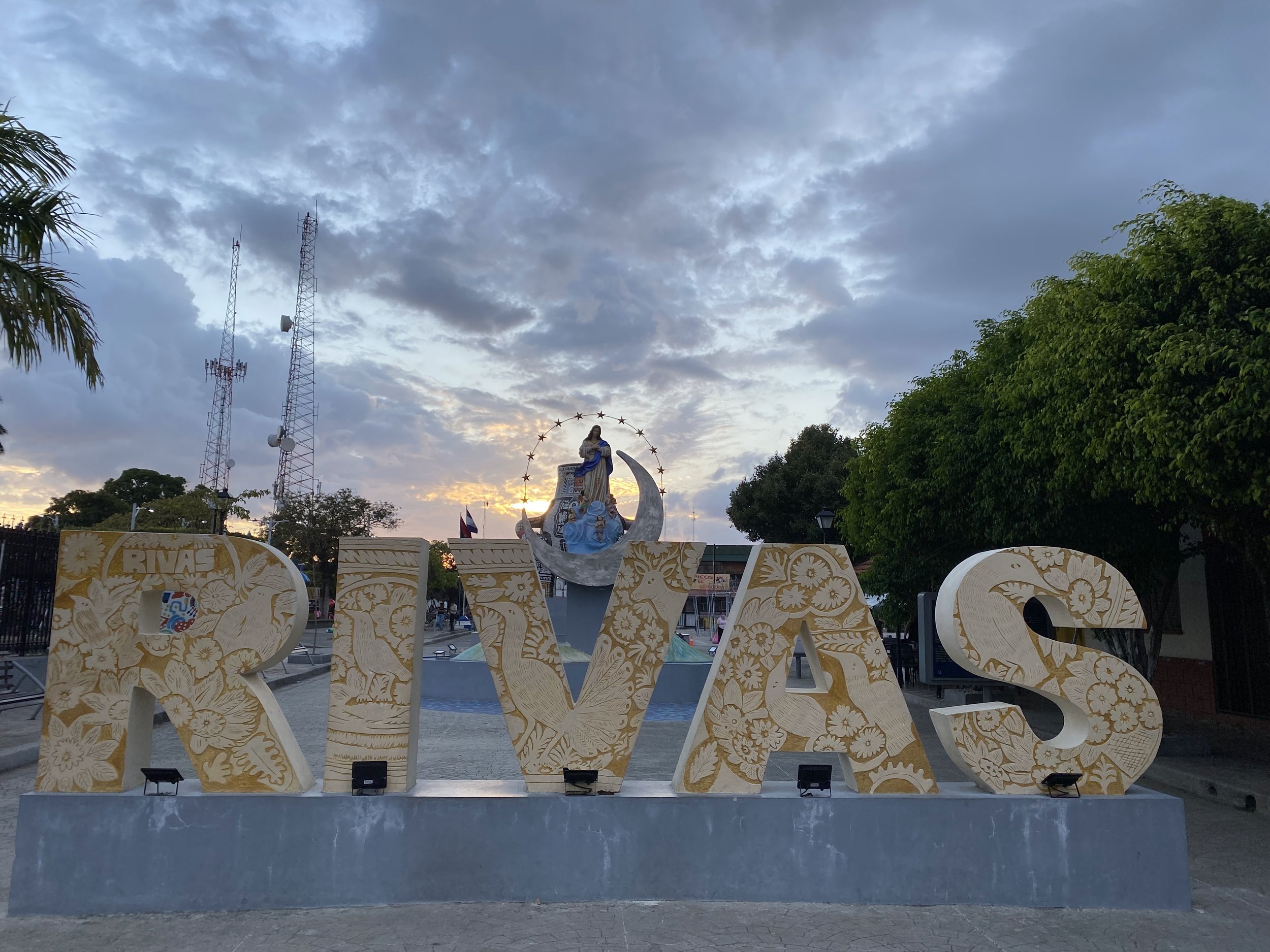 Rivas town sign at sunset in Nicaragua, with dramatic storm clouds overhead and a religious statue behind the large decorative letters in the central square.