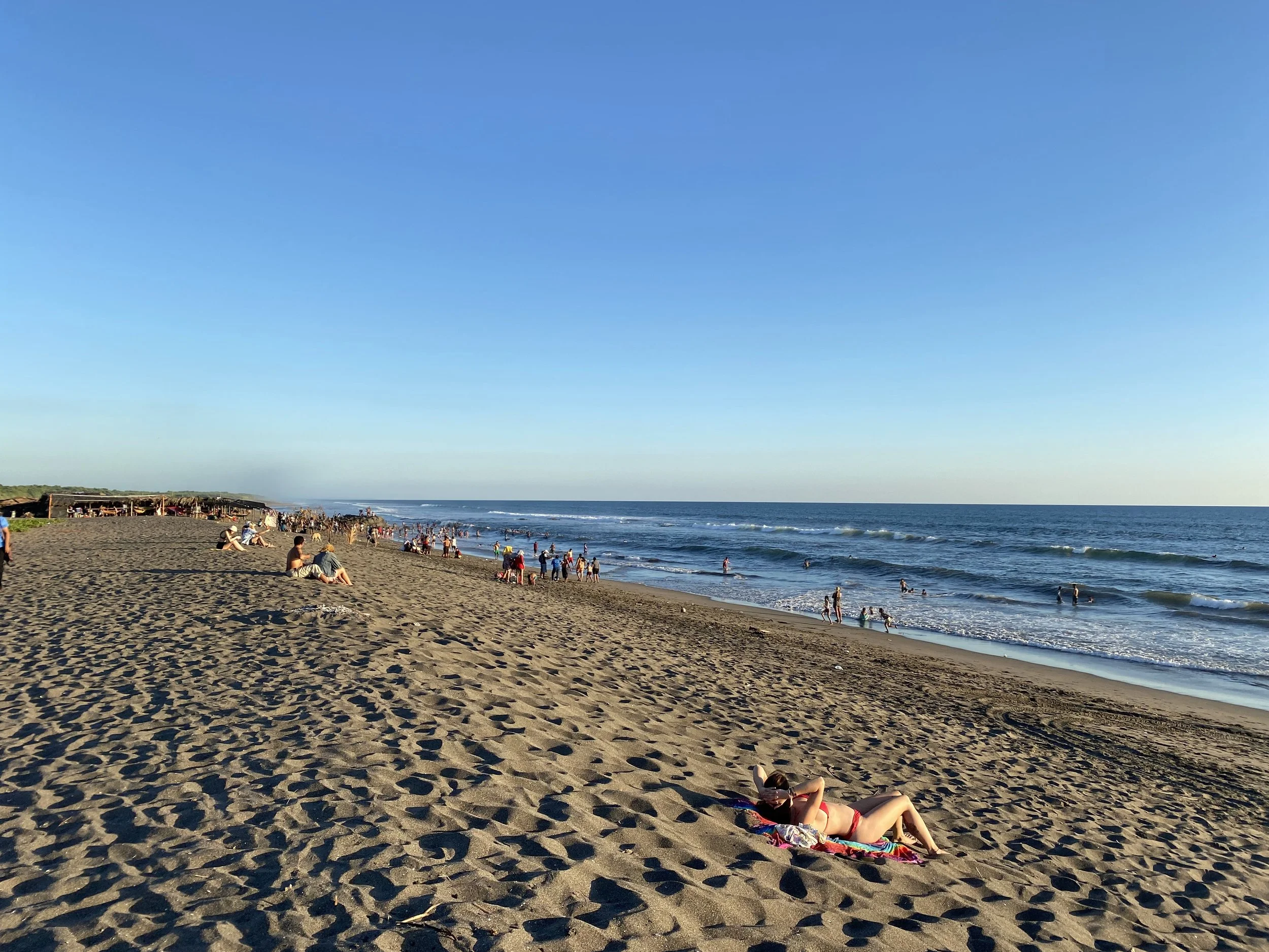 Wide sandy beach in Las Peñitas with people walking, relaxing, and swimming along the shoreline under a clear blue sky.