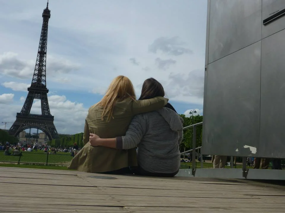 Two friends sitting together with their arms around each other while looking toward the Eiffel Tower in Paris, France.