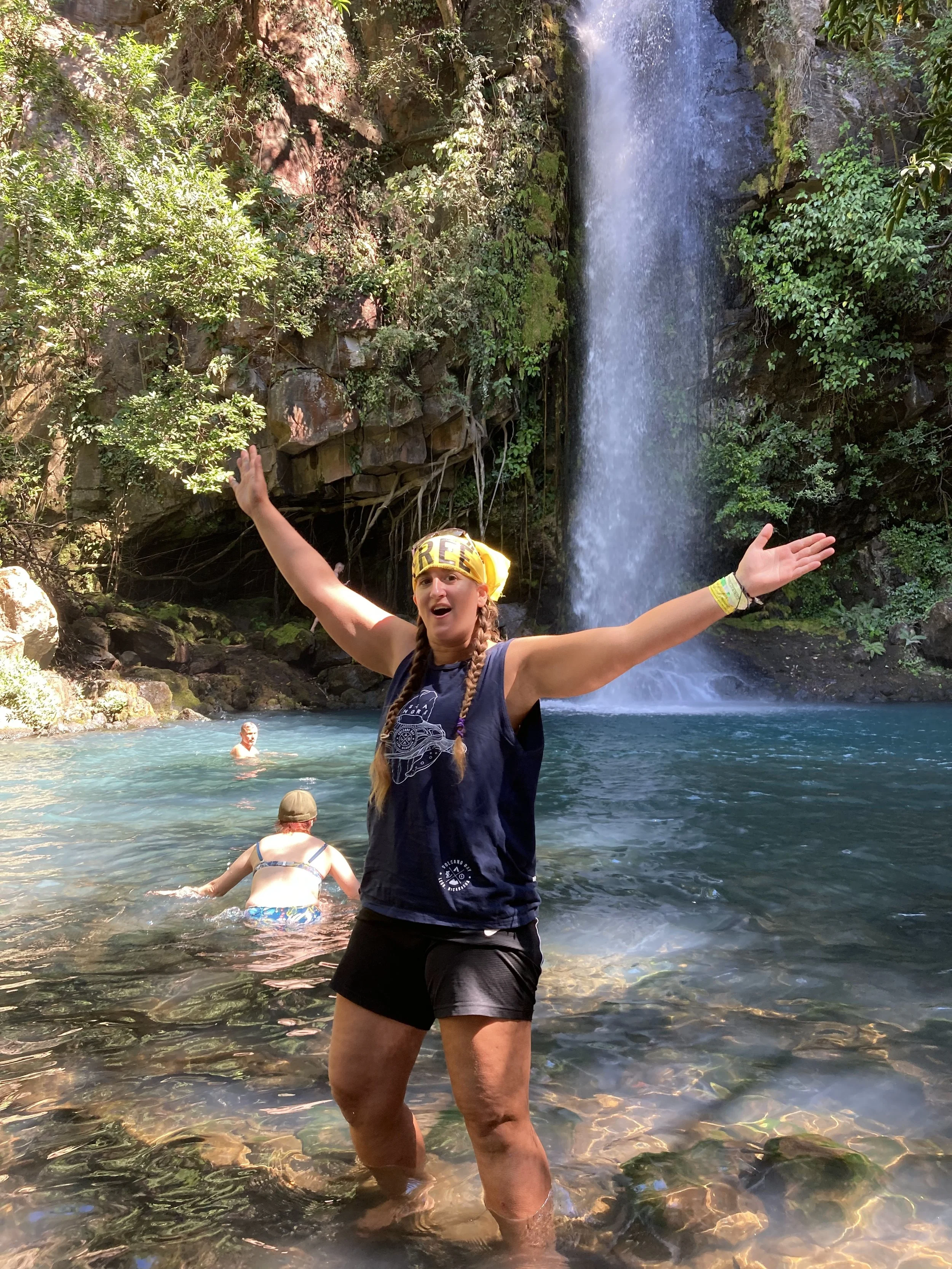 A woman standing in a clear turquoise pool with her arms raised in front of a waterfall at Rincón de la Vieja National Park in Costa Rica, while other visitors swim nearby.