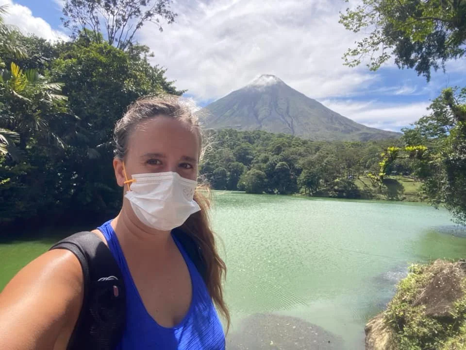 Woman standing beside a green volcanic lake with Arenal Volcano rising in the background in La Fortuna, Costa Rica.