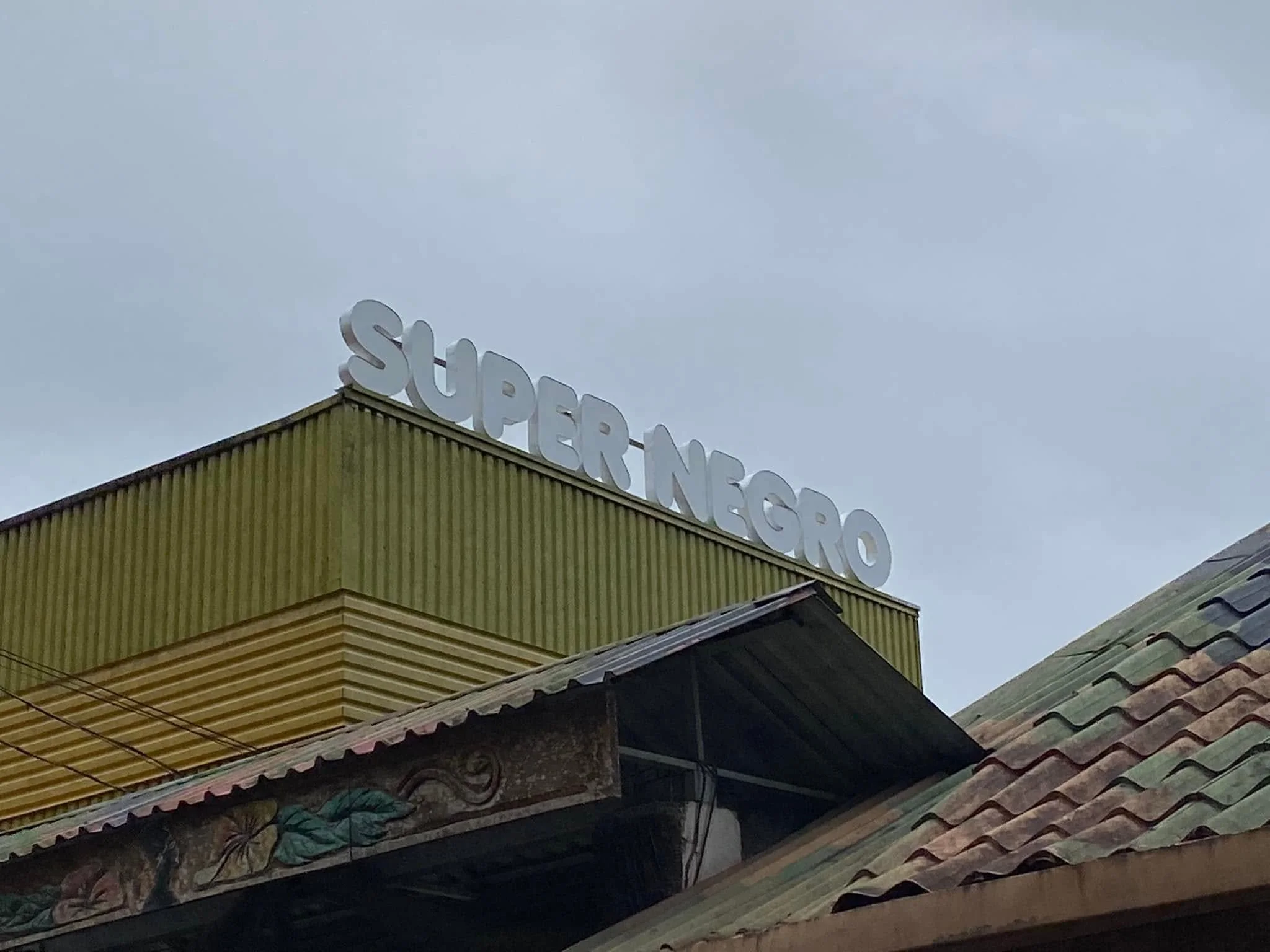 Large white letters spelling “Super Negro” on top of a green corrugated building beneath a cloudy sky in Puerto Viejo.