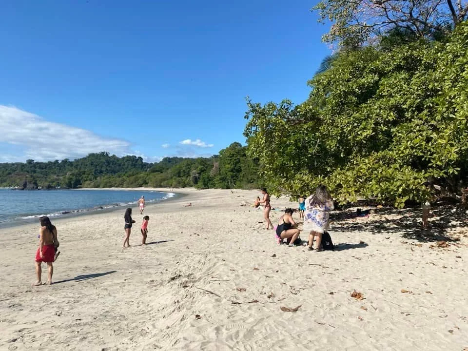 Wide sandy beach inside Manuel Antonio National Park with a few visitors, calm ocean water, and dense green rainforest along the shoreline.