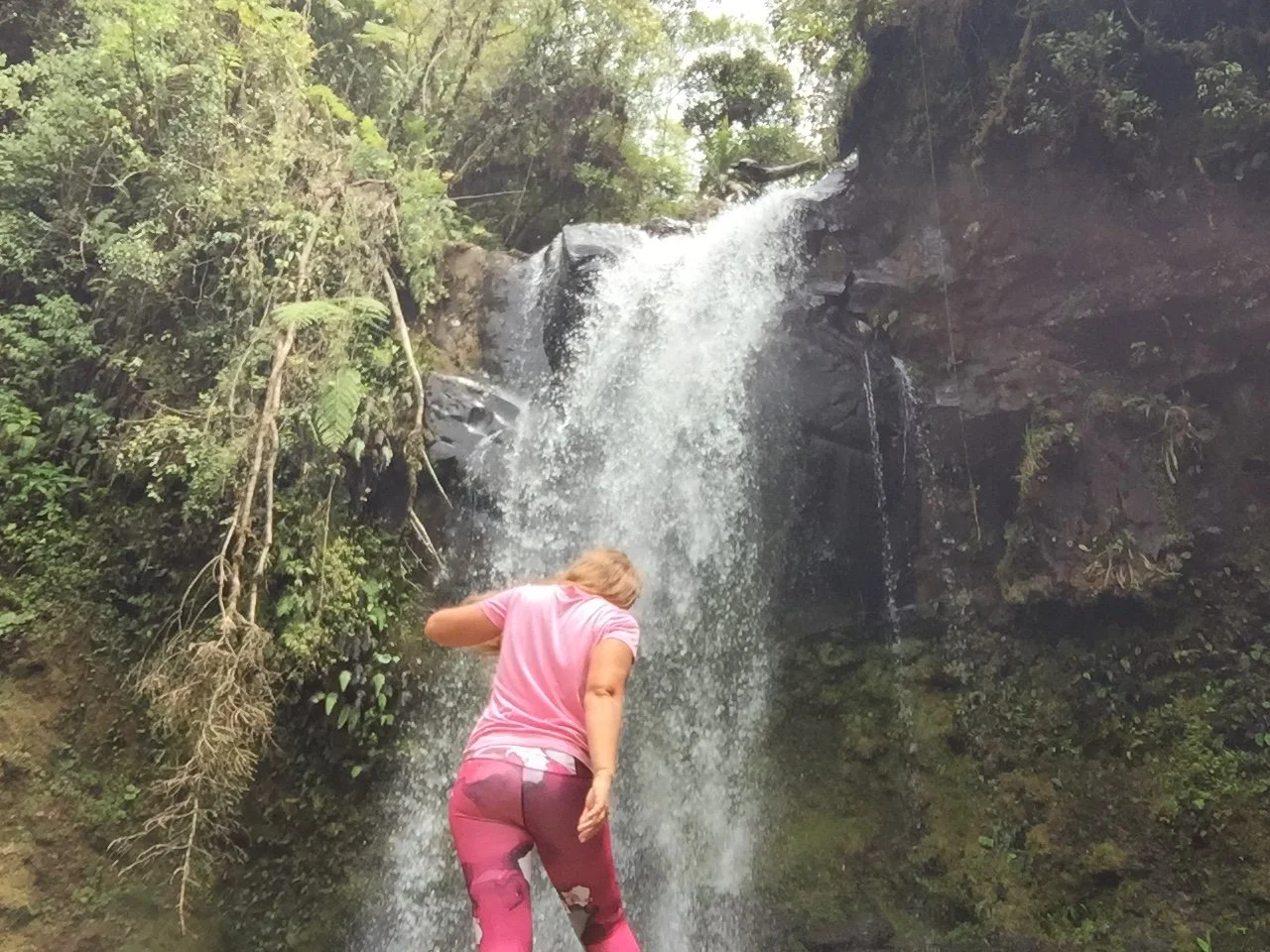 A woman wearing pink hiking clothes climbing over rocks toward a waterfall surrounded by dense green forest in Boquete, Panama.