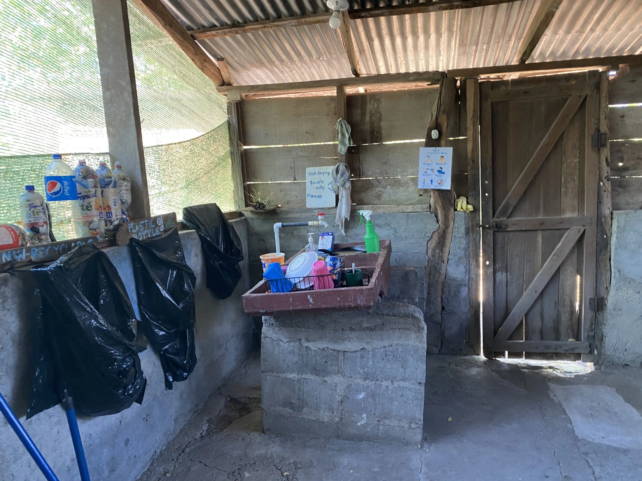 Rustic kitchen washing area at the Cabuyal sea turtle conservation camp, with a concrete sink, dish rack, recycling bags and wooden walls.
