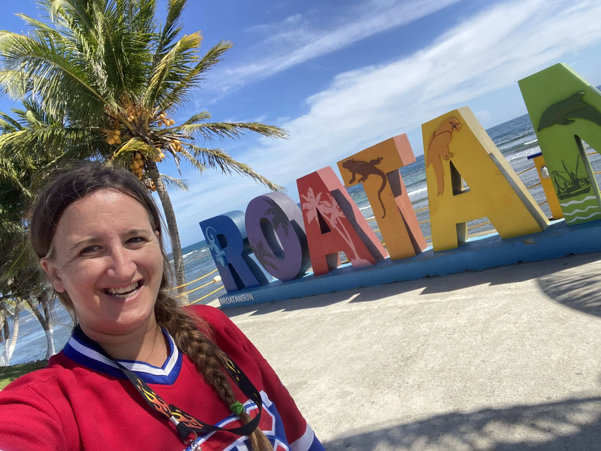 Woman smiling in front of colorful Roatán sign with palm trees and ocean in the background in Honduras