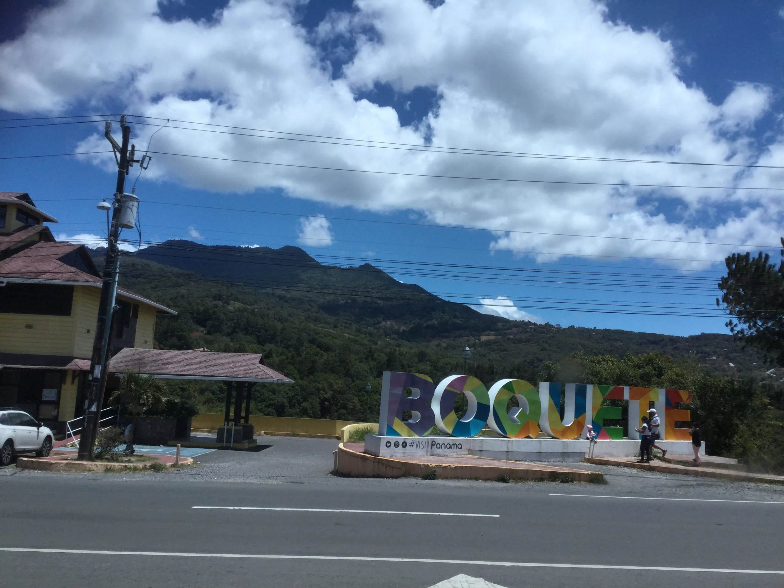 The colourful Boquete sign beside a road, with green mountains and a partly cloudy blue sky in the background.
