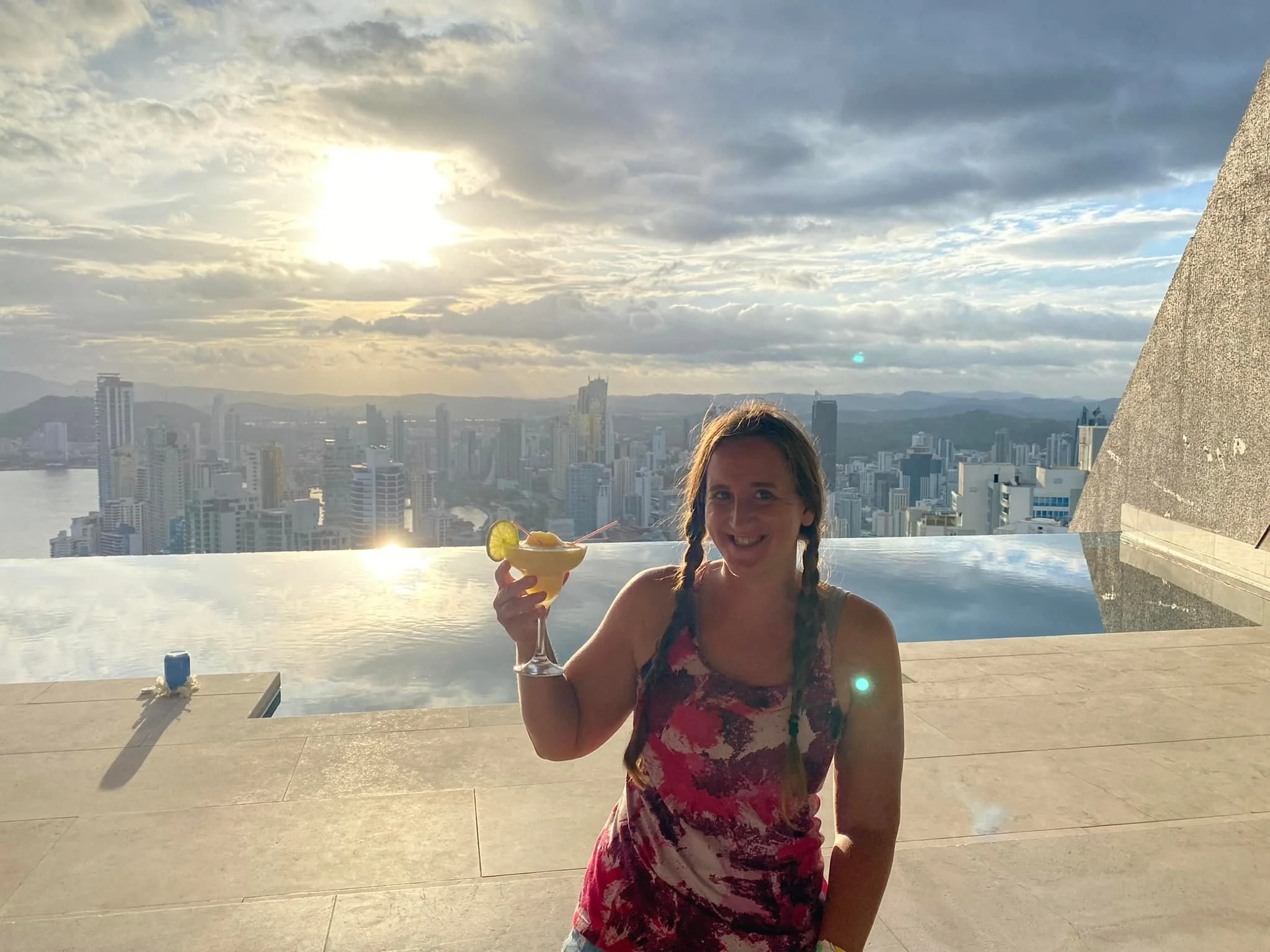 Solo female traveller holding a cocktail beside a rooftop infinity pool at sunset overlooking the Panama City skyline.