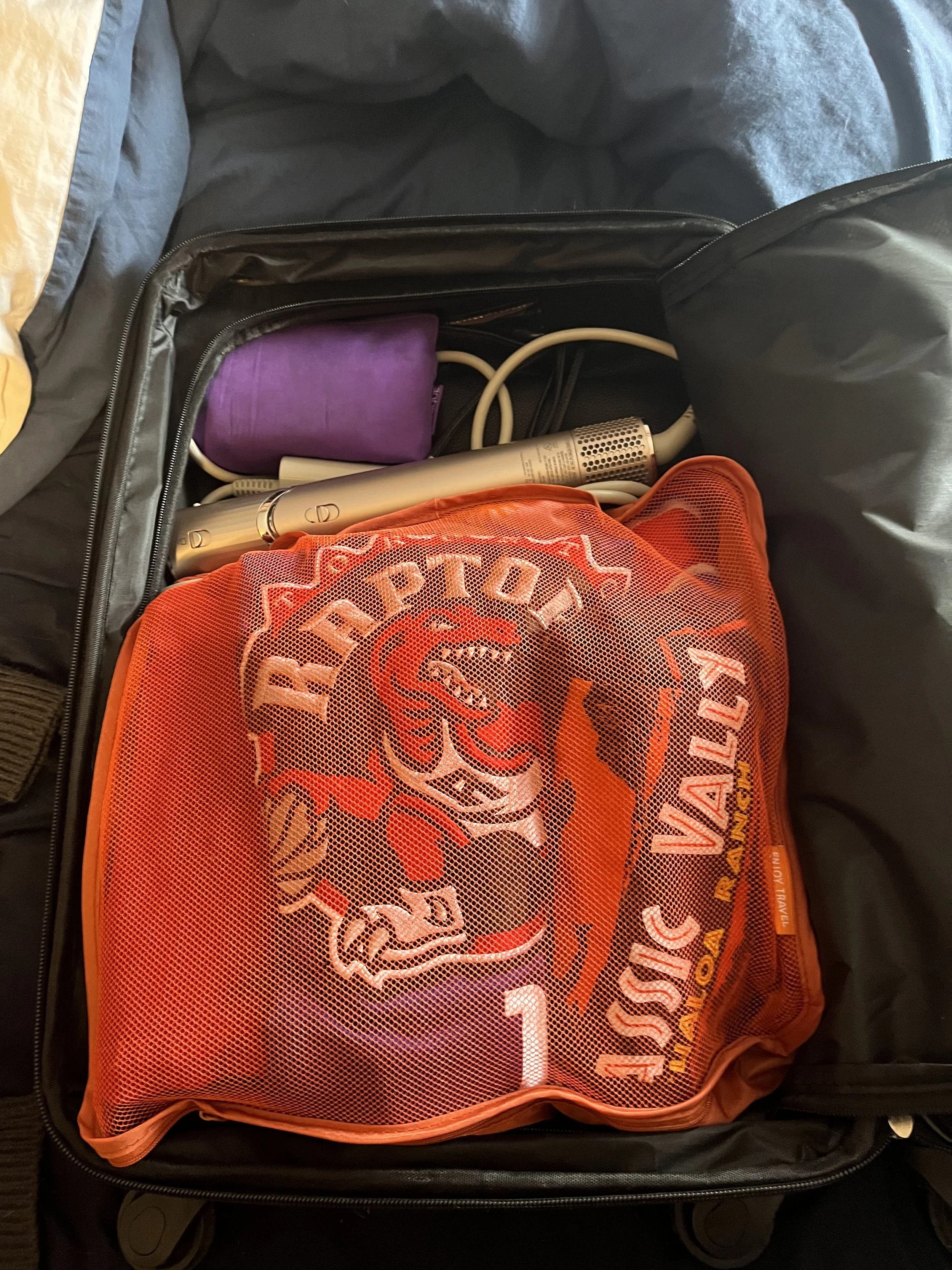 Open suitcase with an orange packing cube filled with clothes, shoes and hair styling tools packed for a trip.