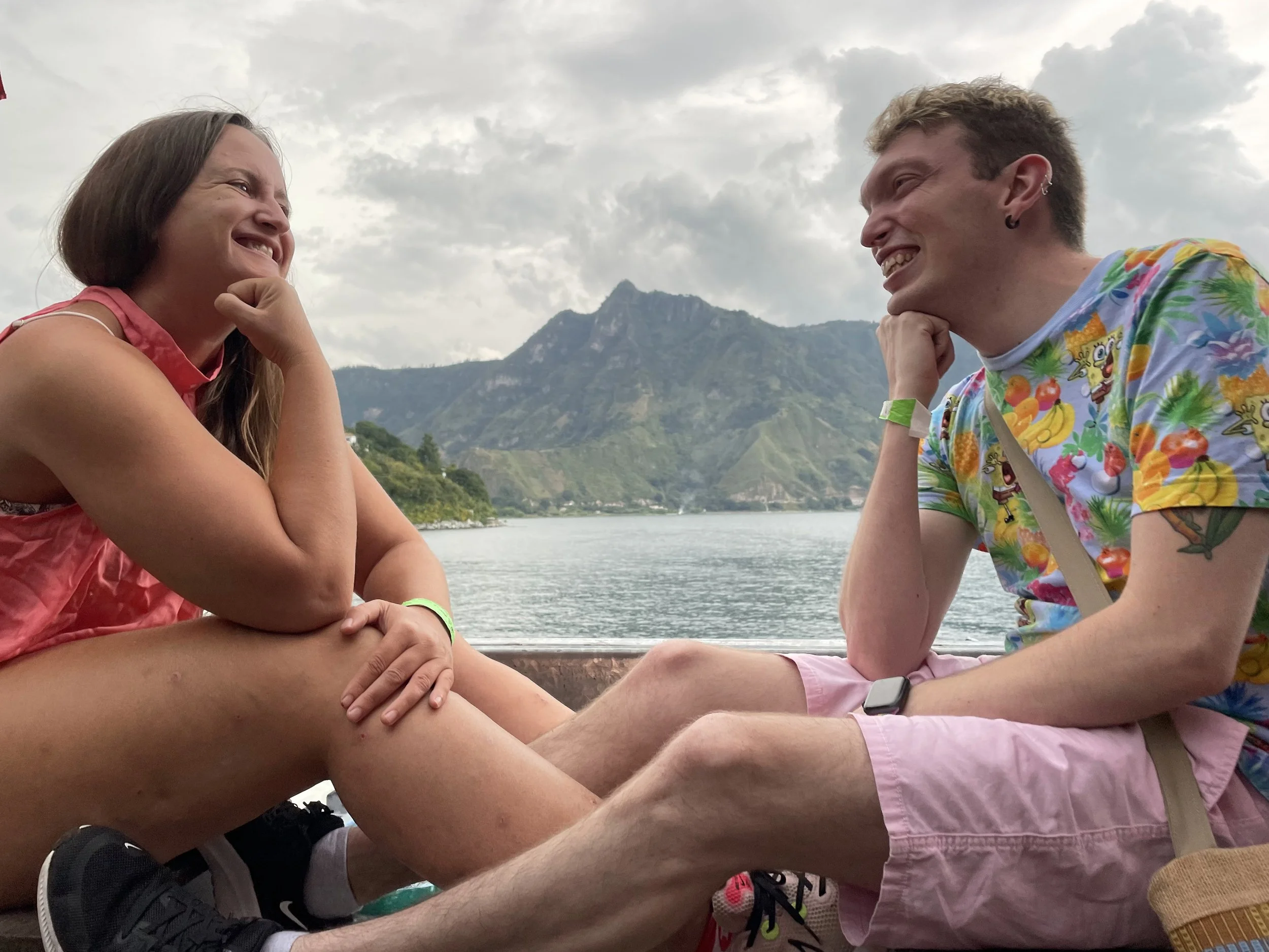 Two friends smiling at each other while sitting on a patio overlooking Lake Atitlán, with mountains and water in the background.
