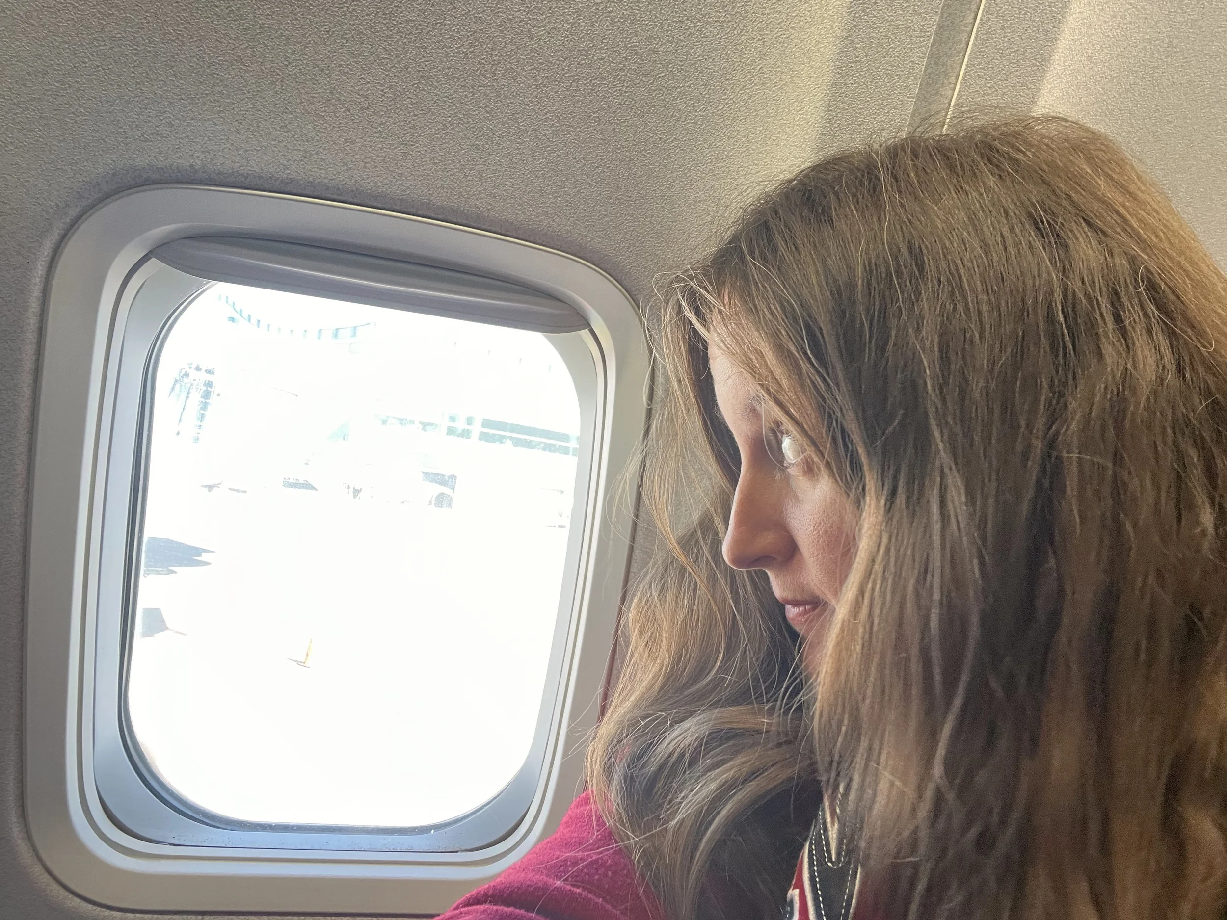 A female traveller sitting beside an airplane window before a flight, looking out toward the airport runway.