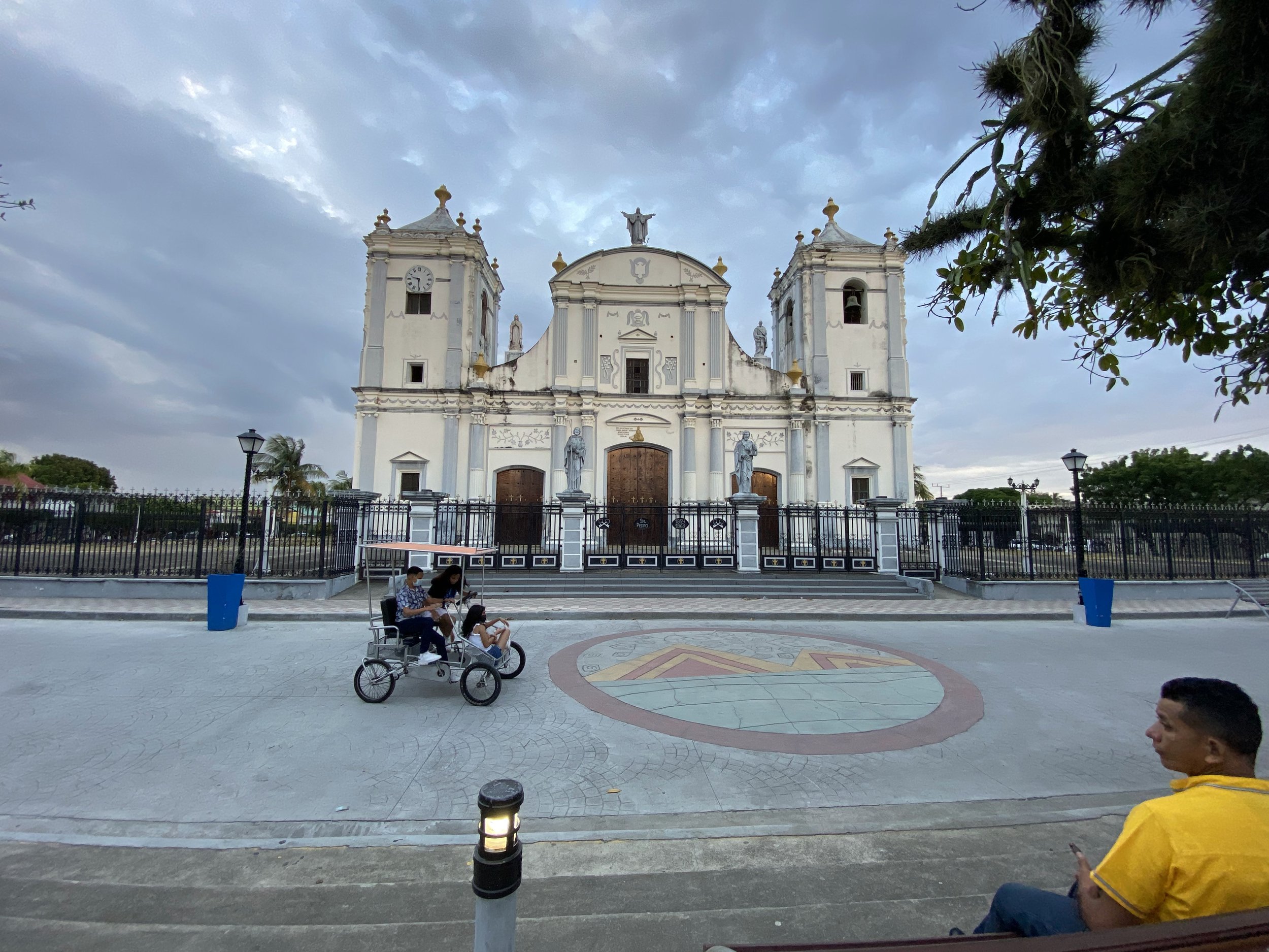 Large white cathedral in the center of Rivas, Nicaragua, under dark storm clouds at sunset, with locals riding through the square on a bicycle cart.