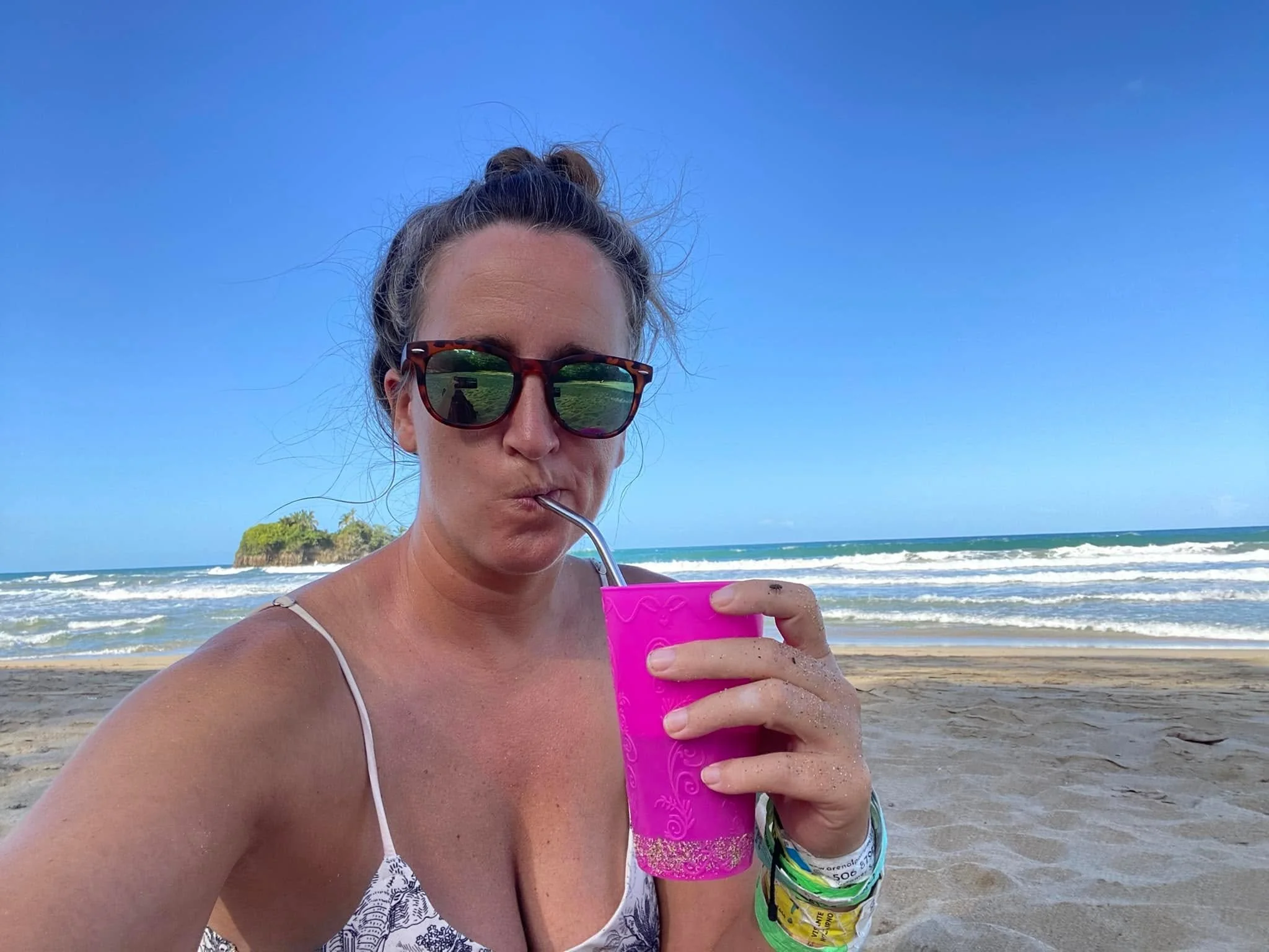 Woman wearing sunglasses drinking from a bright pink cup while relaxing on a sandy beach in Puerto Viejo, Costa Rica, with waves and the Caribbean Sea behind her.