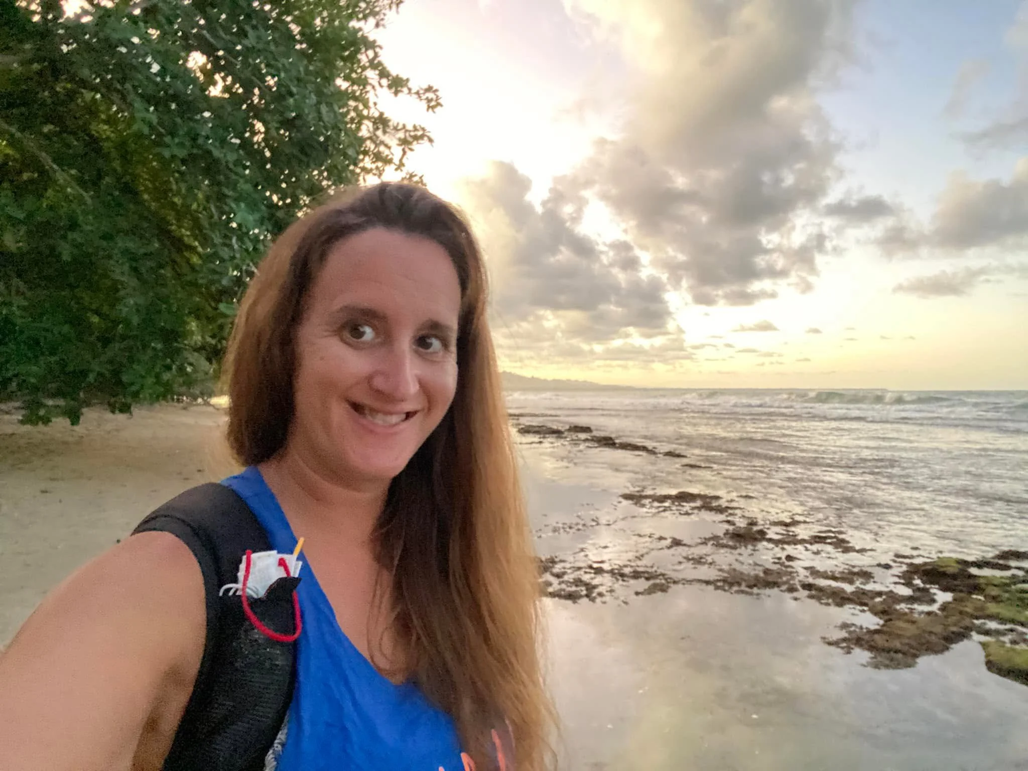 Woman taking a selfie while walking along a sandy beach in Puerto Viejo, Costa Rica, with the Caribbean Sea and a cloudy sunset in the background.