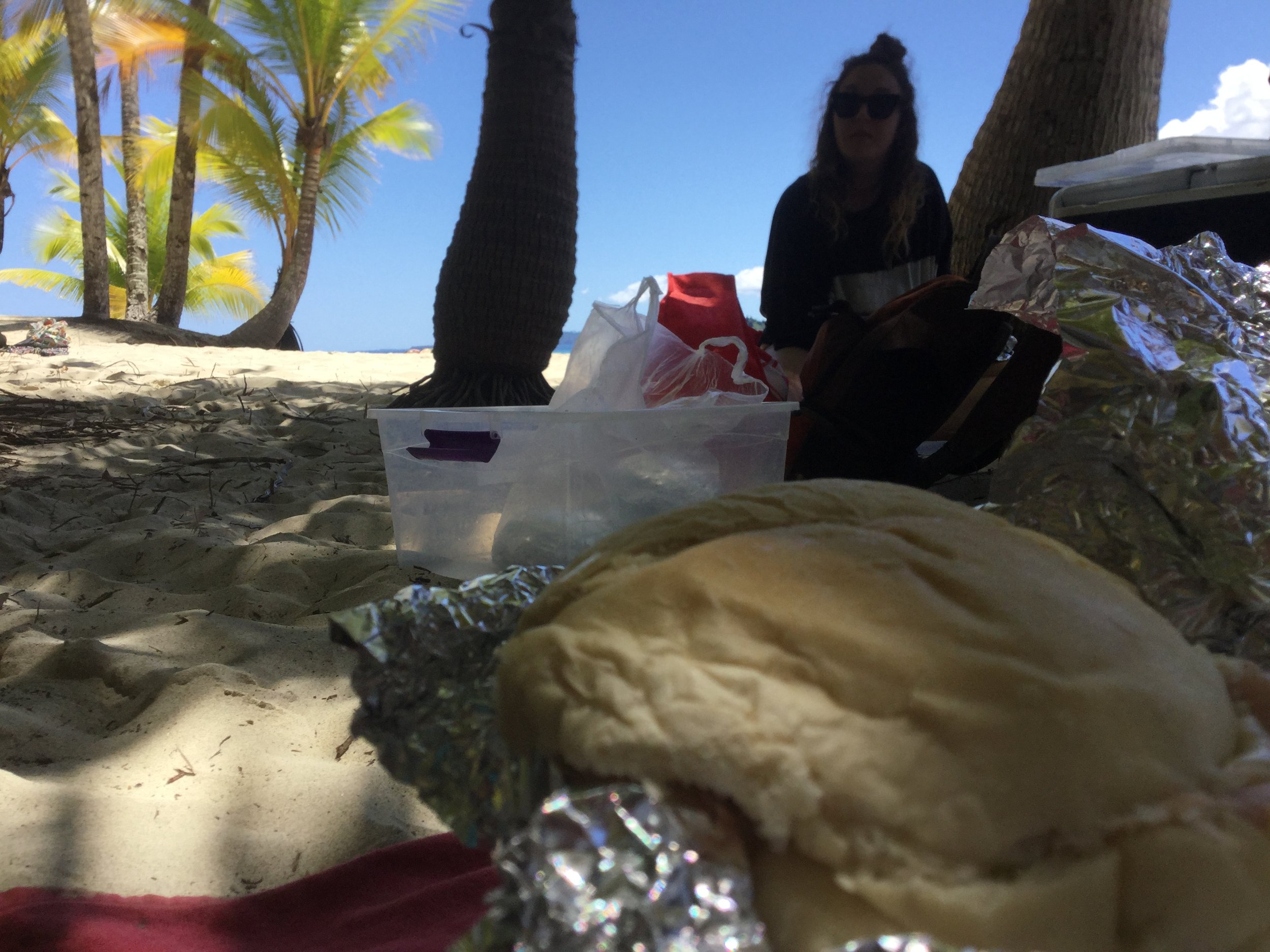Foil-wrapped sandwich in the foreground with a woman sitting beneath palm trees on a sandy beach.
