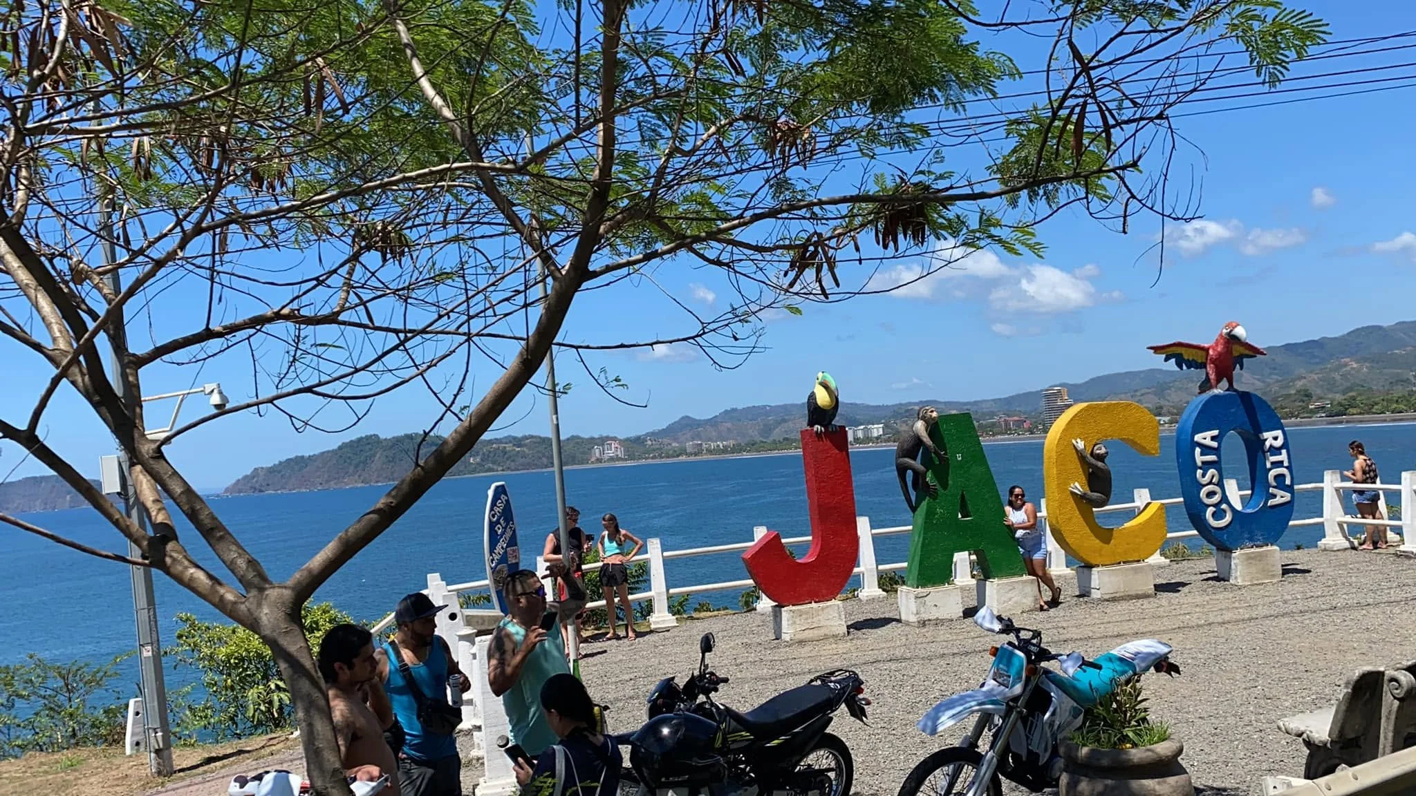 The colourful "JACO" sign overlooking the Pacific Ocean in Jacó, Costa Rica, with visitors enjoying the waterfront promenade on a sunny day.