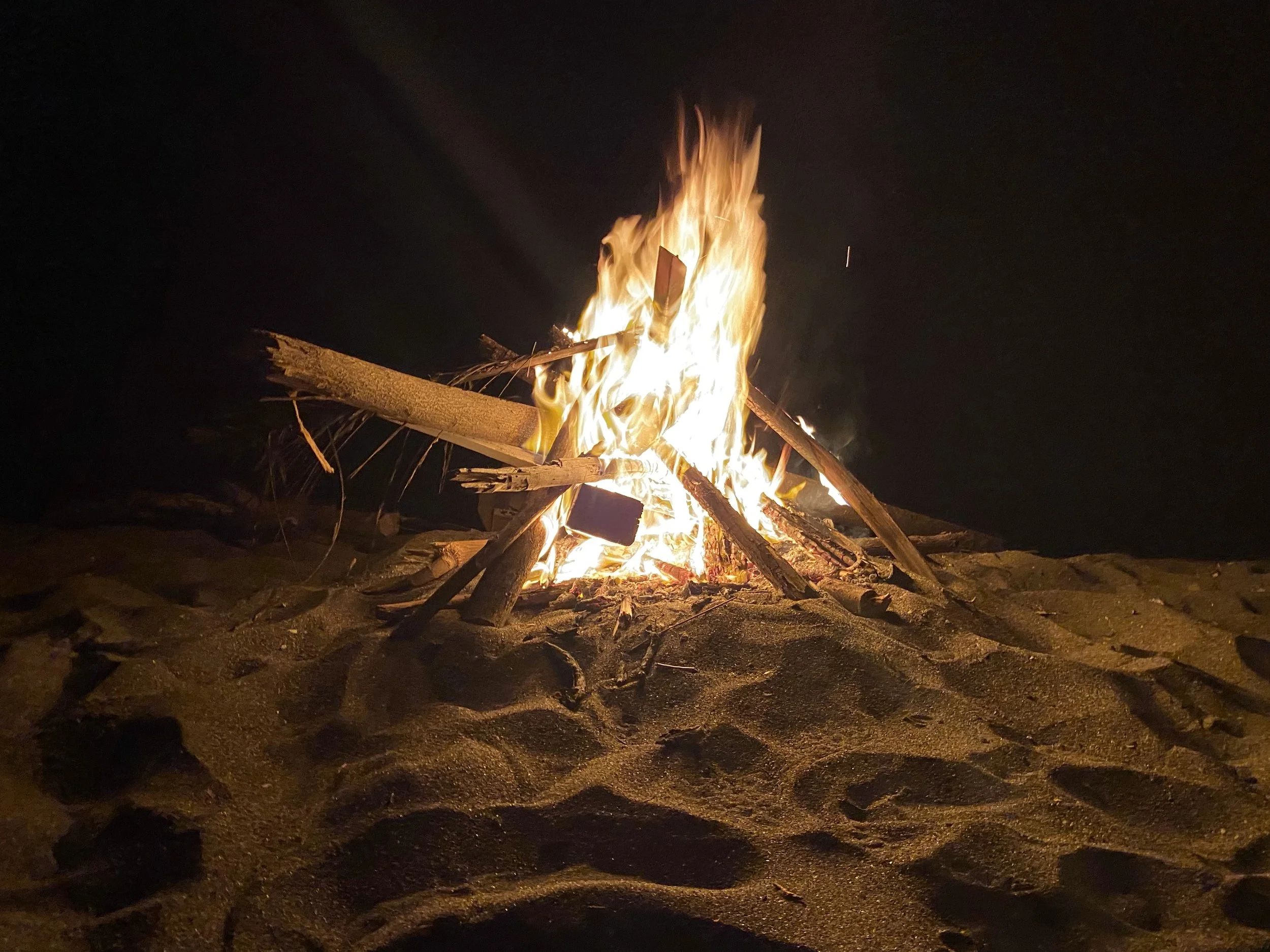 Beach bonfire burning on the sand at night in Las Peñitas, surrounded by darkness with flames lighting the scene.