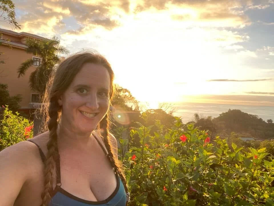 A woman with braided hair smiling beside a roadside garden as the sun sets over the Pacific Ocean in Manuel Antonio, Costa Rica.