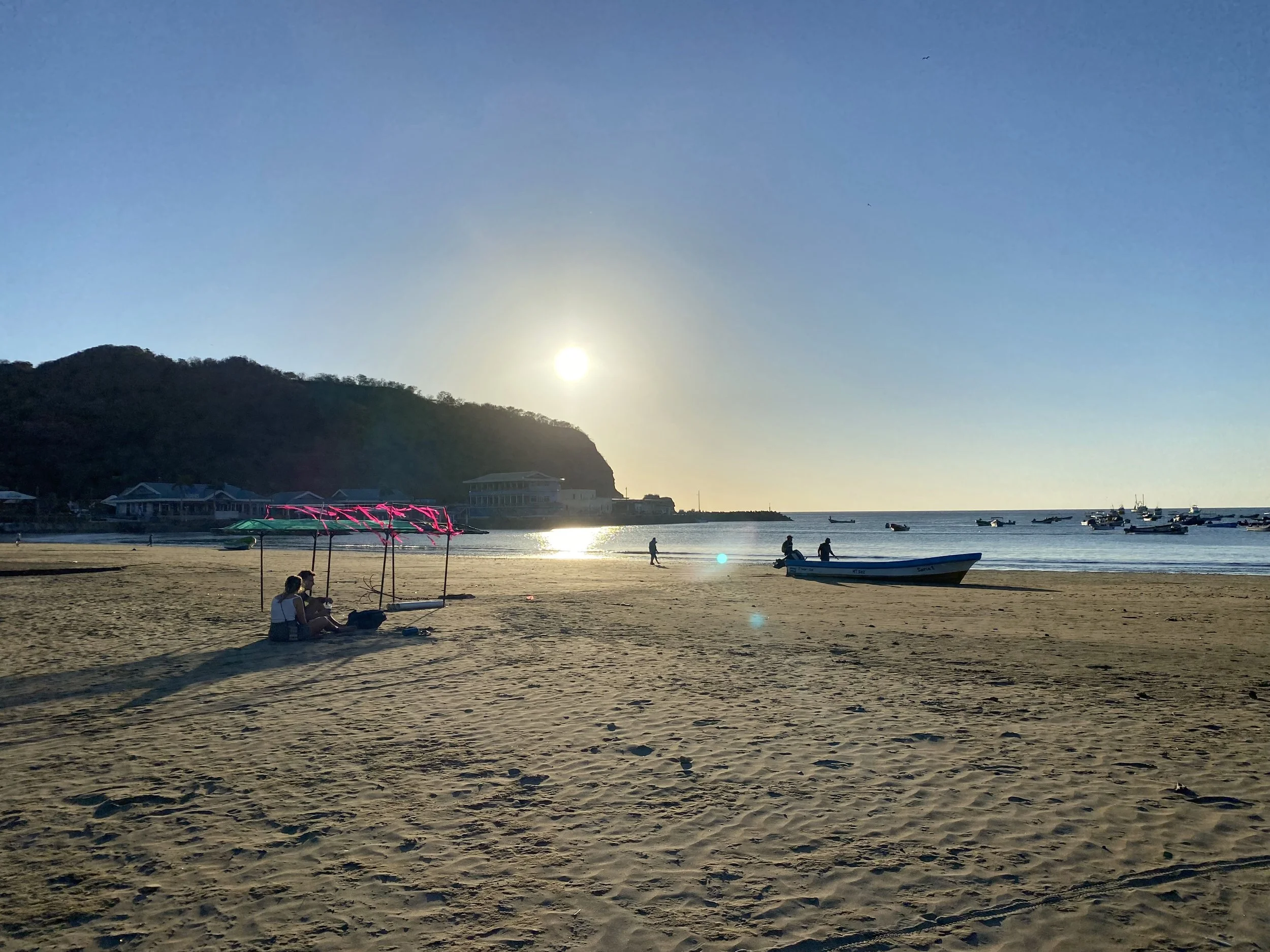 Sunset over the beach in San Juan del Sur, Nicaragua, with boats anchored in the bay and people relaxing on the sand.