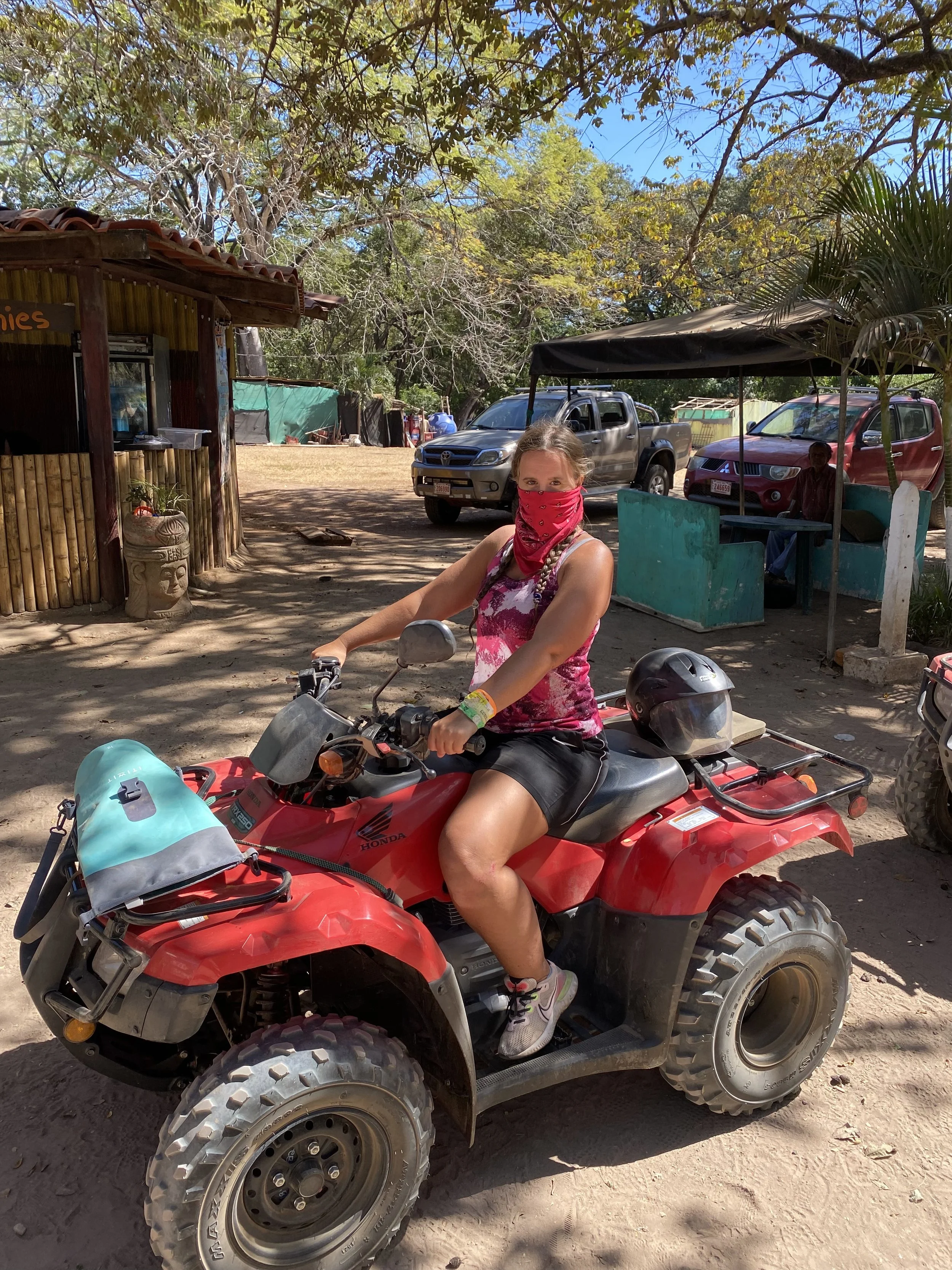 A woman sitting on a red ATV before the start of an off-road tour near Tamarindo, Costa Rica, wearing a bandana over her face and surrounded by tropical scenery.