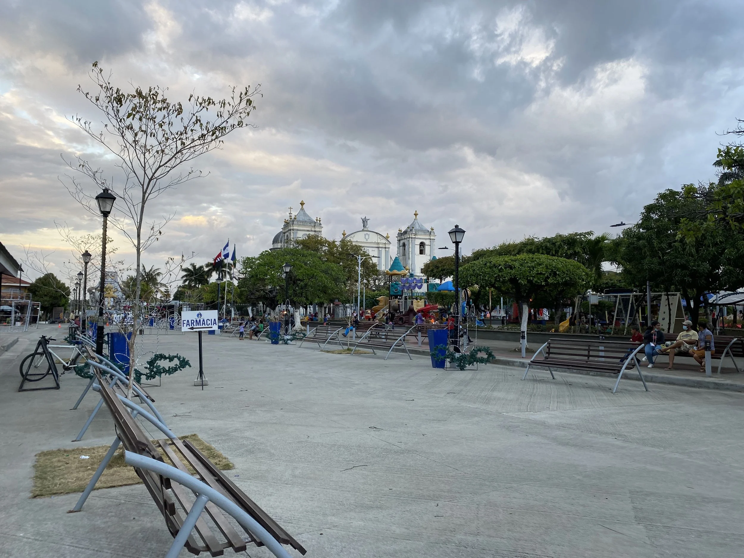 Quiet town square in Rivas, Nicaragua, with benches, locals relaxing, storm clouds overhead, and the cathedral visible in the background at sunset.