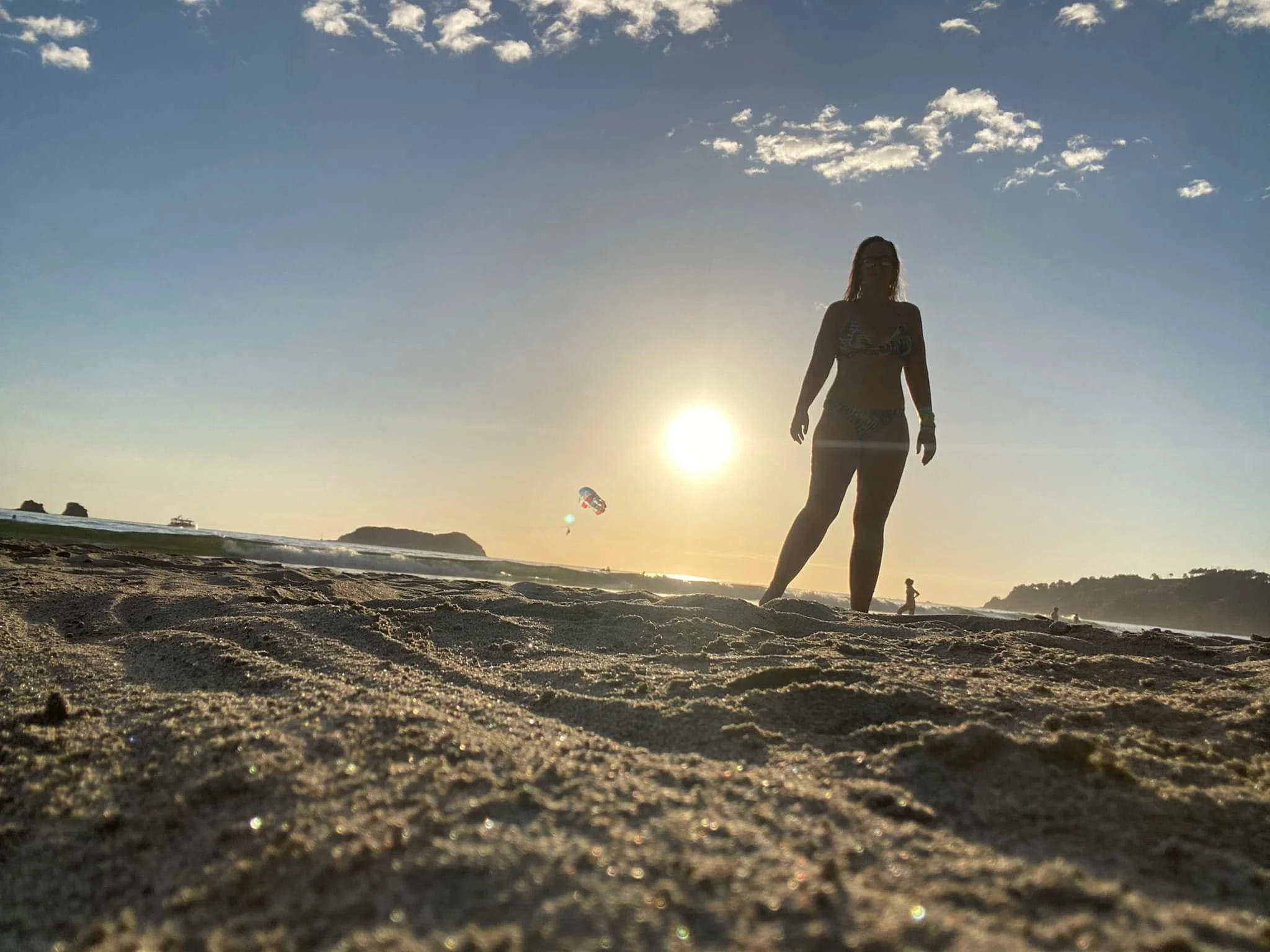 Woman standing on Playa Espadilla at sunset with the Pacific Ocean, parasailing, and the sun setting behind her in Manuel Antonio, Costa Rica.