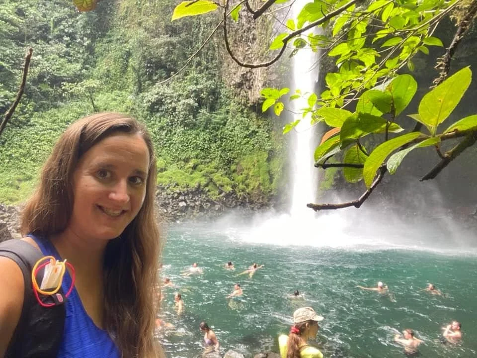 La Fortuna Waterfall cascading into a turquoise swimming hole surrounded by rainforest, with visitors enjoying the water and a traveller standing in the foreground.