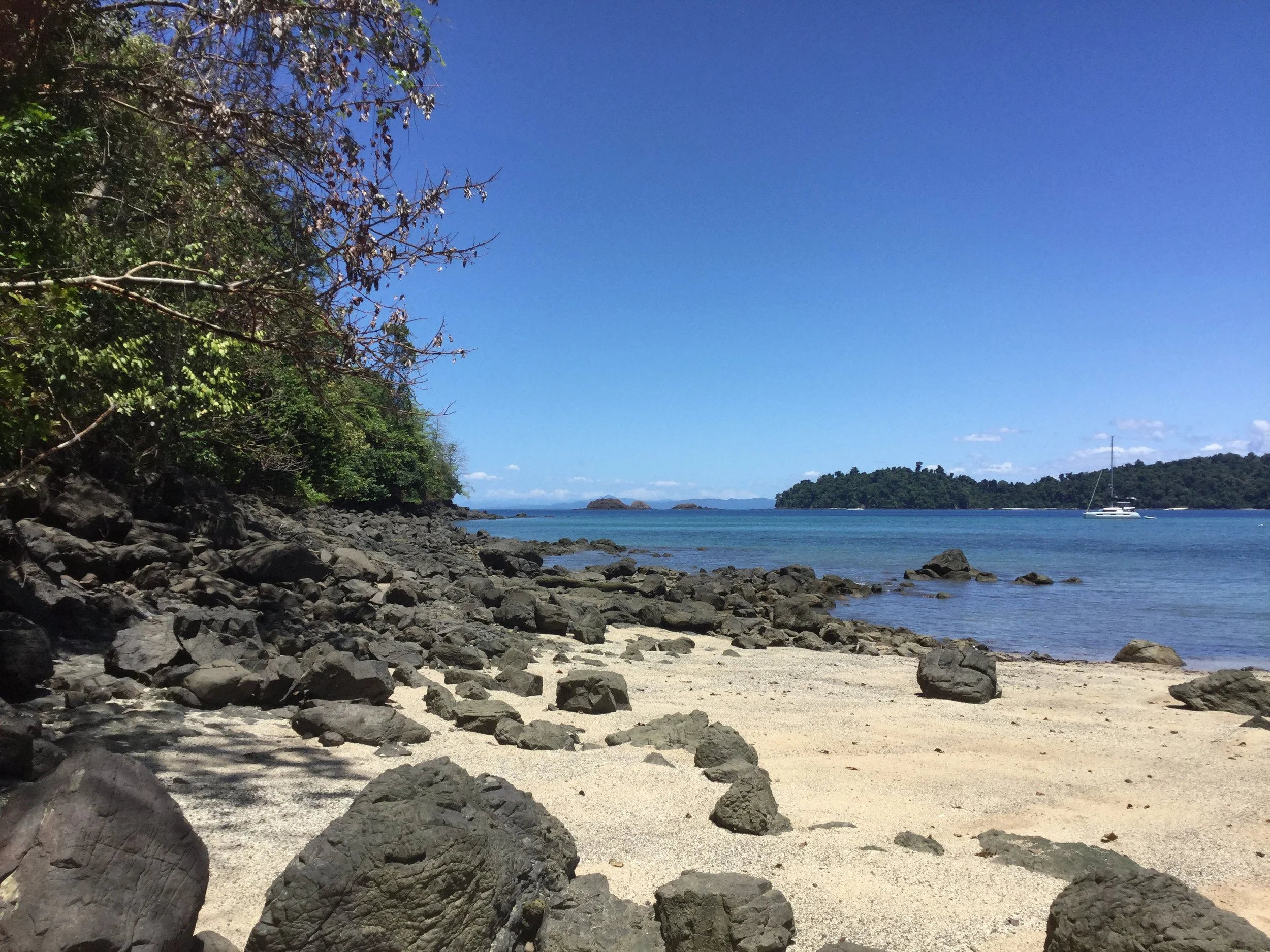 Rocky tropical shoreline beside calm blue water, with forested islands and a white sailboat in the distance.