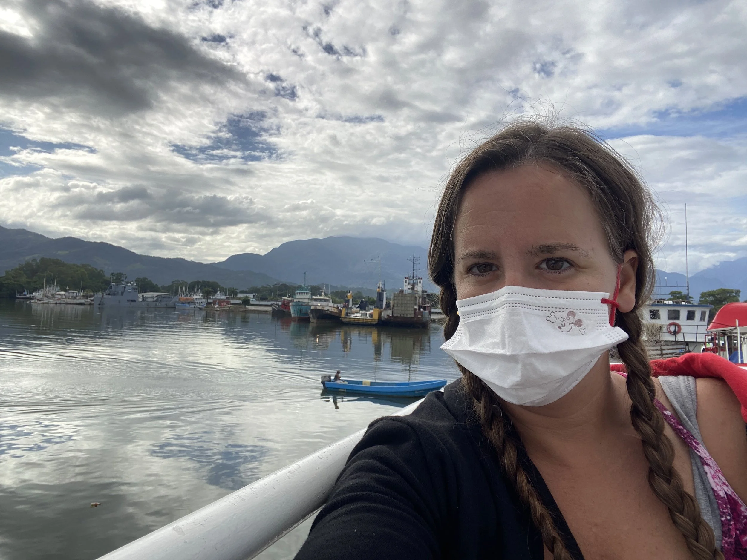 Woman wearing a face mask on the top deck of the ferry leaving mainland Honduras for Utila, with boats and mountains behind her.