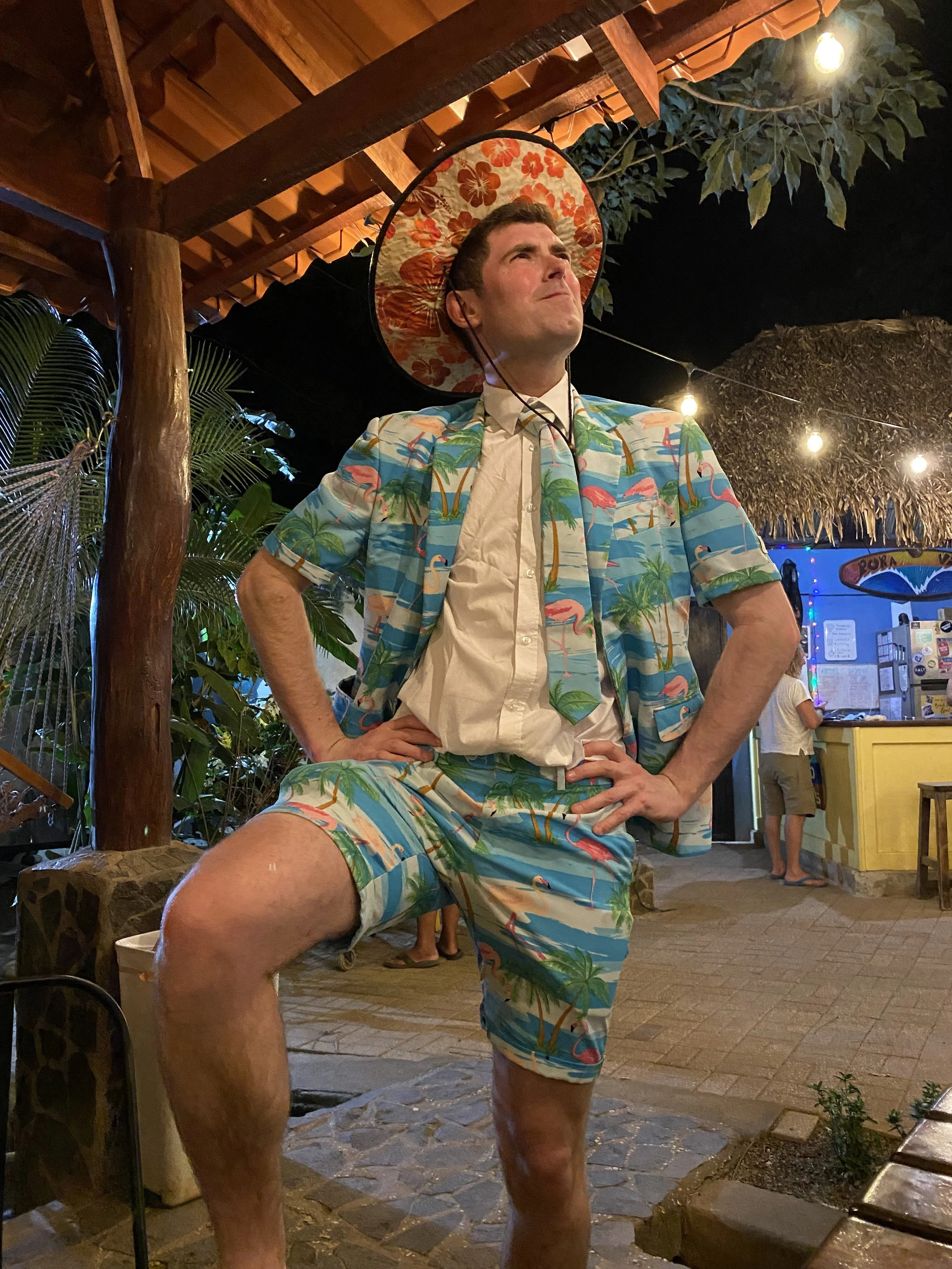 A traveller wearing a colourful tropical outfit and sun hat poses in the common area of a backpacker hostel in Tamarindo, Costa Rica during an evening social gathering.