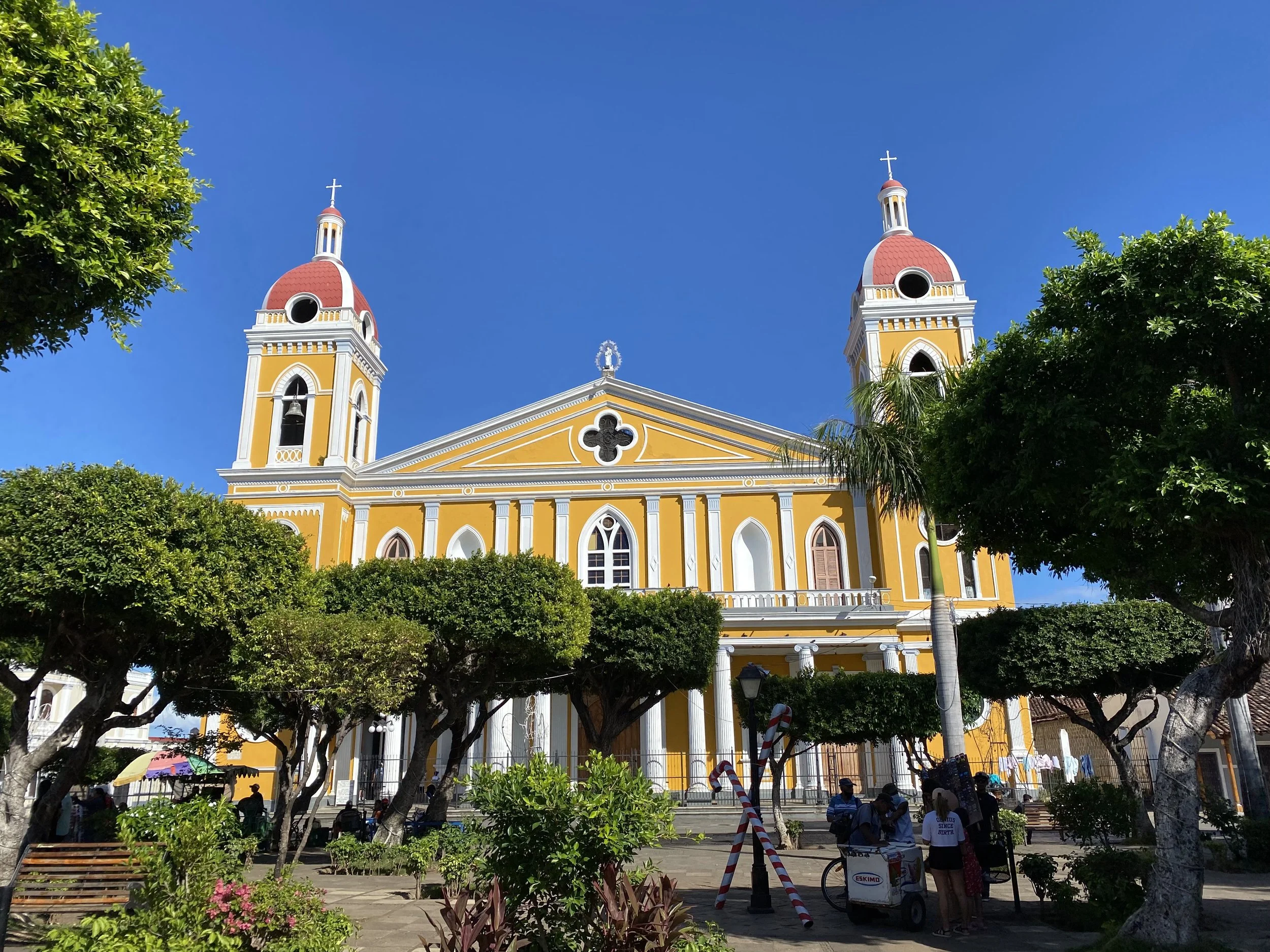 Granada Cathedral with yellow walls and red domes seen from a plaza with trees and people in the foreground under a bright blue sky.