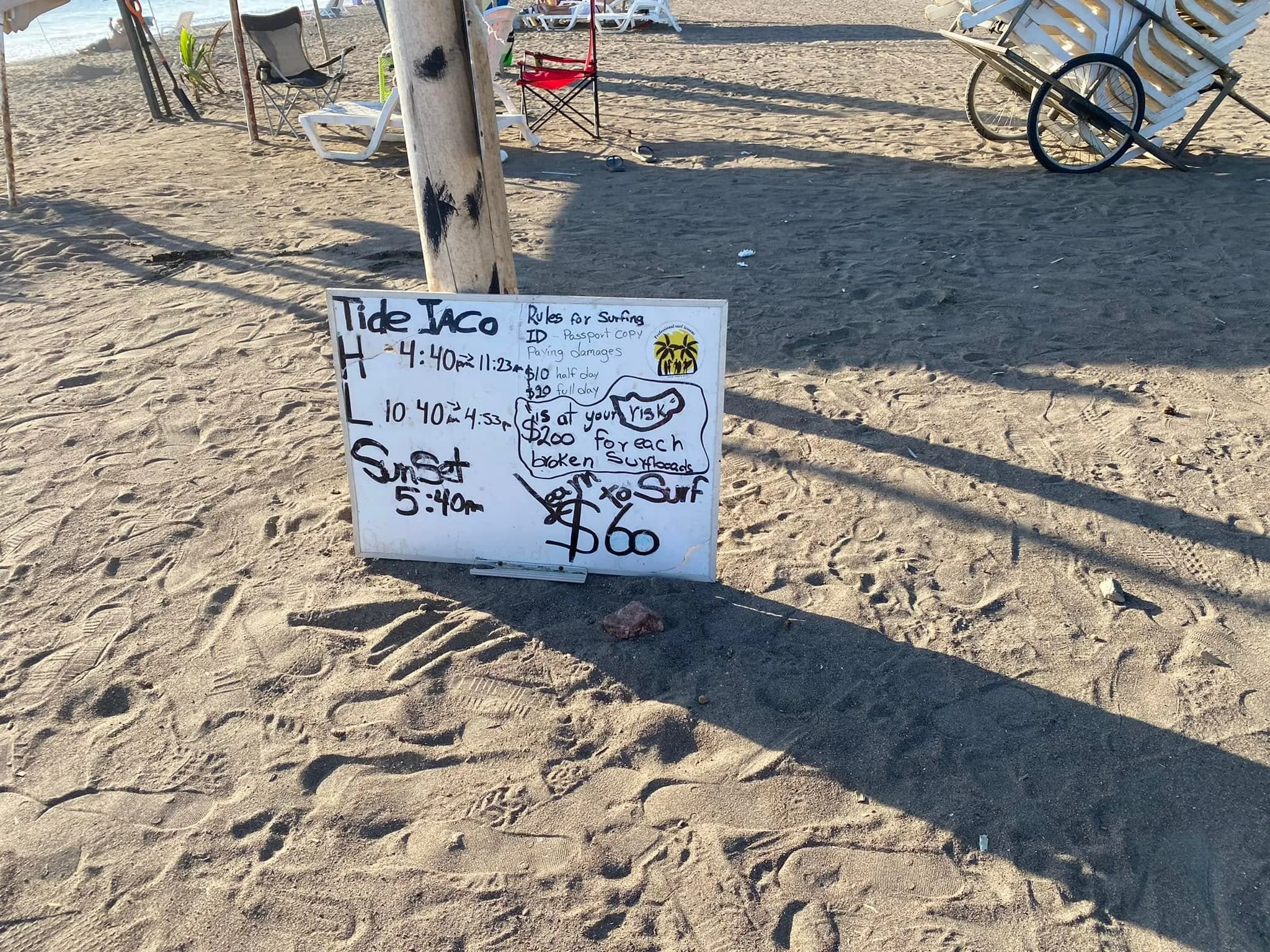 A sign on Jacó Beach in Costa Rica displaying surf lesson information, tide times, and the day's sunset time, with the sandy beach and ocean in the background.