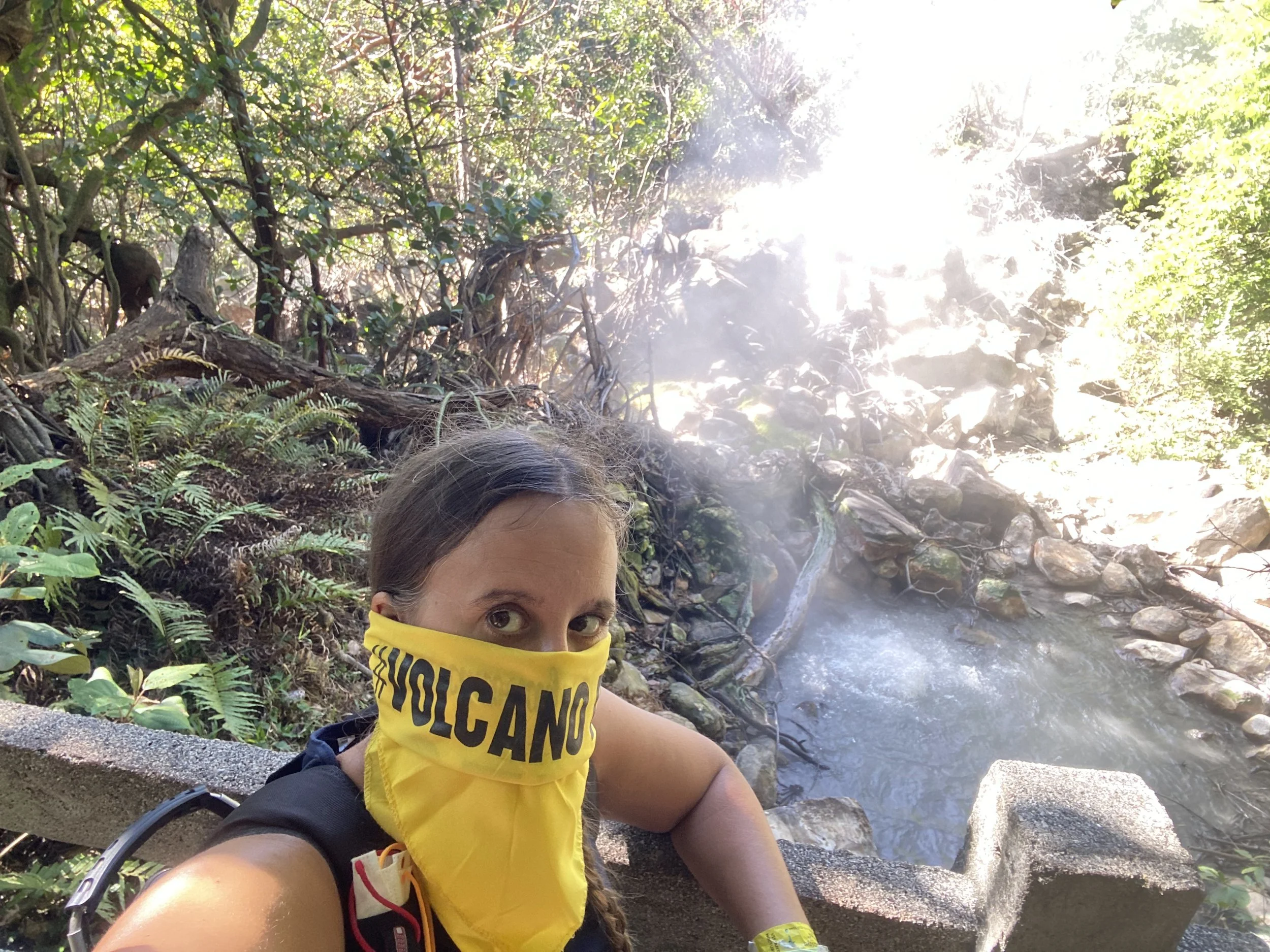 Person wearing a yellow "Volcano" bandana over their nose and mouth beside steaming geothermal pools at Rincón de la Vieja National Park in Costa Rica.