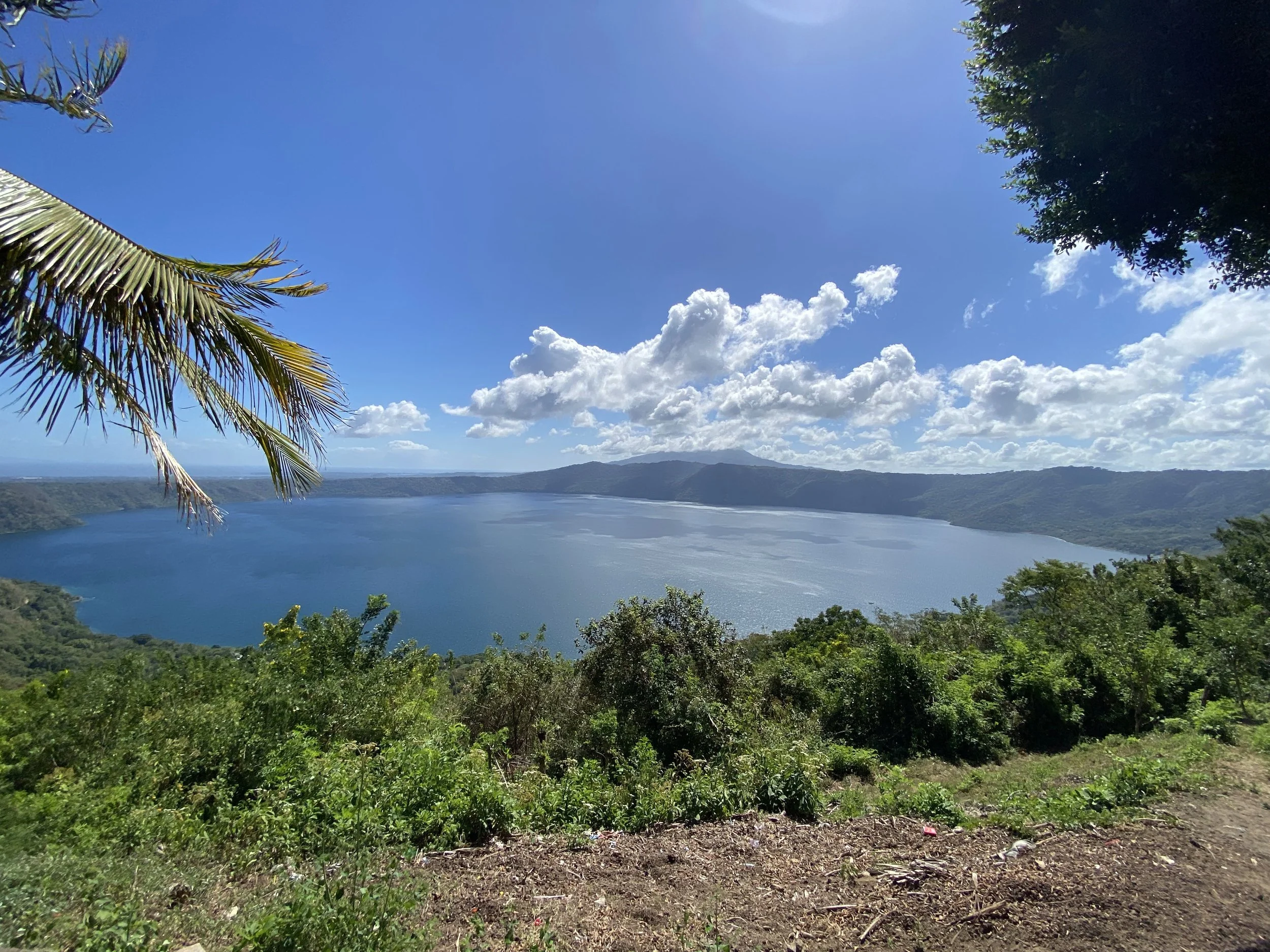 View of Laguna de Apoyo crater lake in Nicaragua, surrounded by green hills and trees under a bright blue sky with scattered clouds.