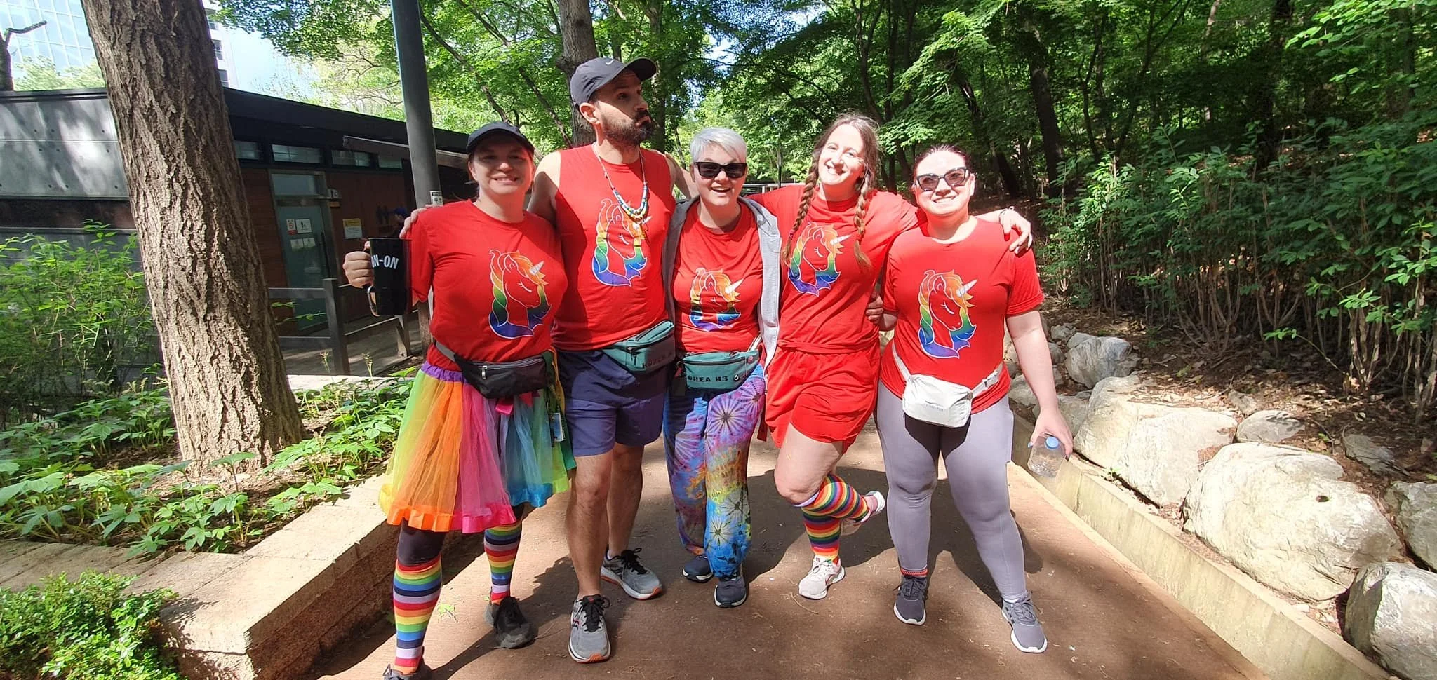 A group of friends wearing red rainbow unicorn shirts and colorful Pride outfits while posing together outdoors.