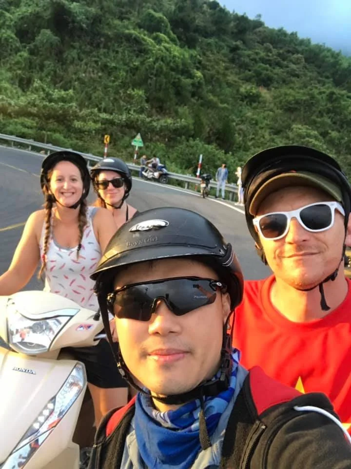 Three travelers and a local friend wearing helmets while exploring Da Nang, Vietnam, on scooters together.