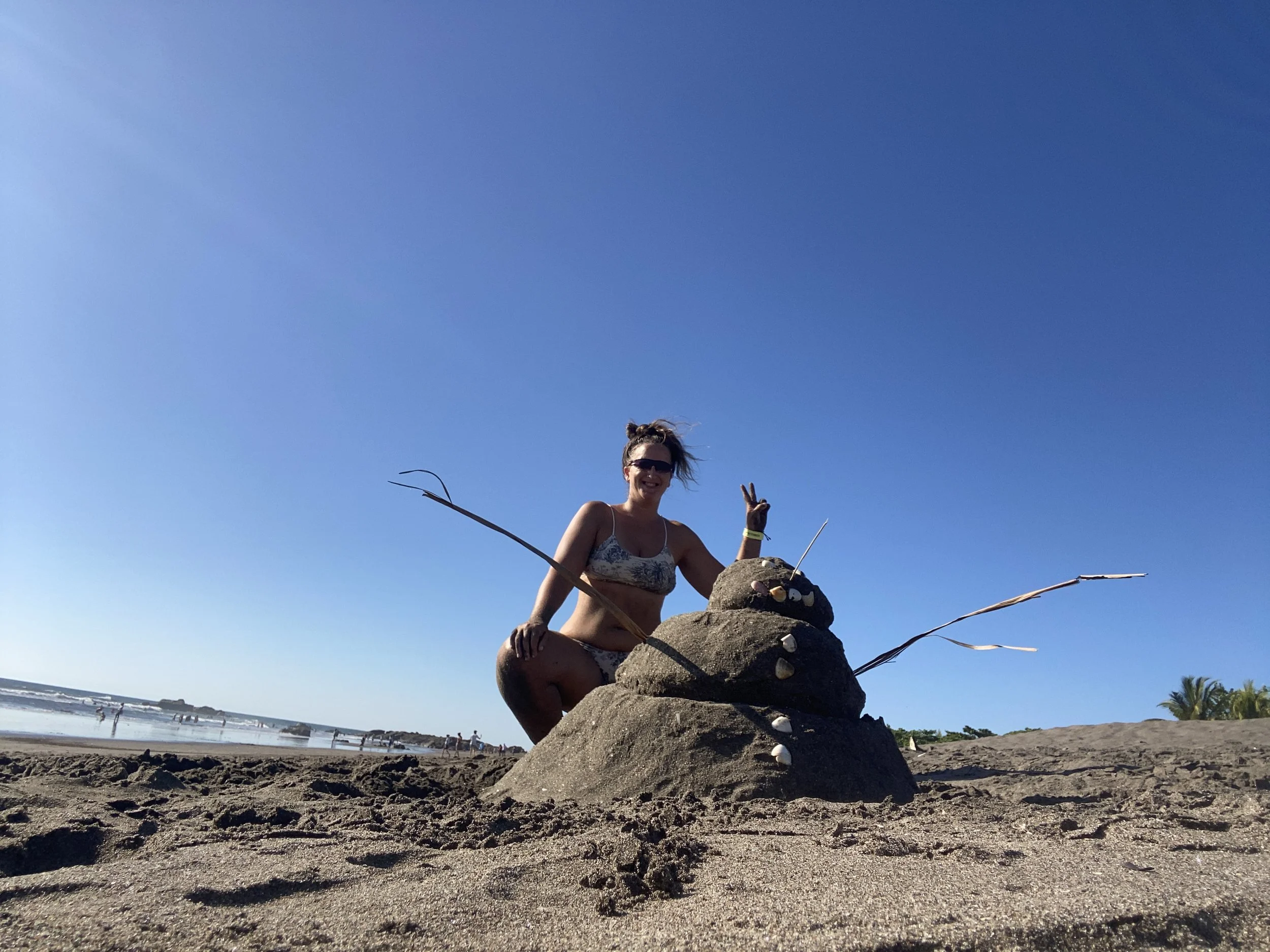 Woman posing beside a sandman sculpture on Las Peñitas beach under a bright blue sky during Christmas in Nicaragua.