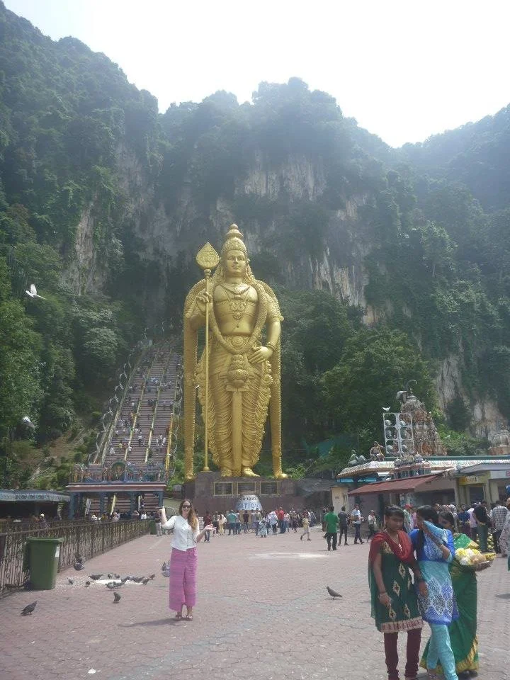 Traveller visiting Batu Caves in Malaysia, with the large golden Murugan statue and colourful staircase rising into the limestone cliffs behind her.