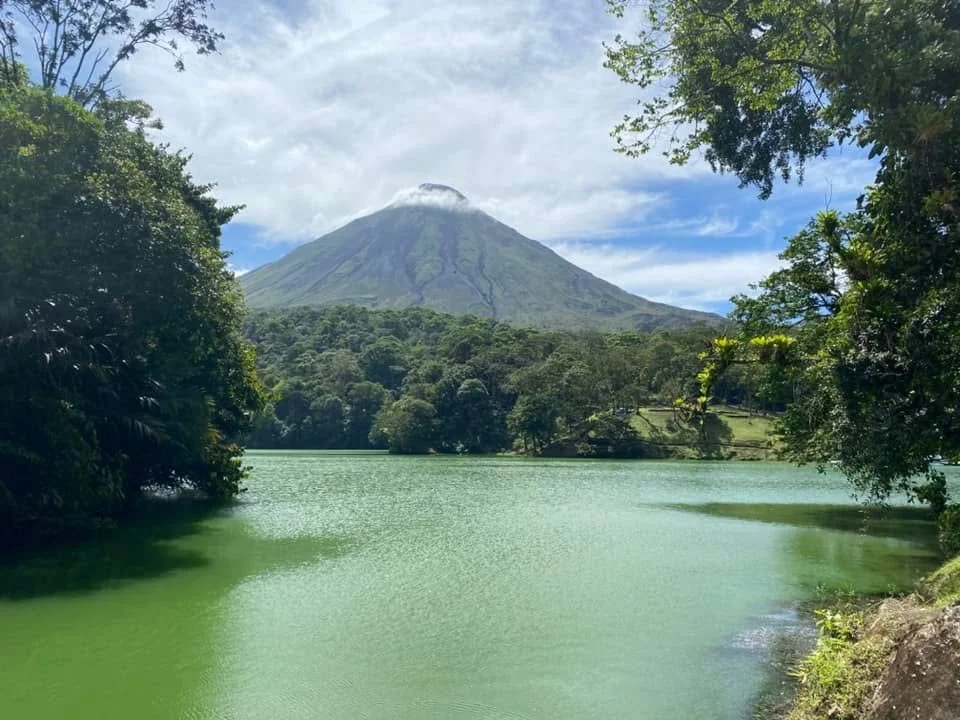 Arenal Volcano rising above a tranquil green lake, surrounded by lush tropical rainforest under a partly cloudy sky in La Fortuna, Costa Rica.