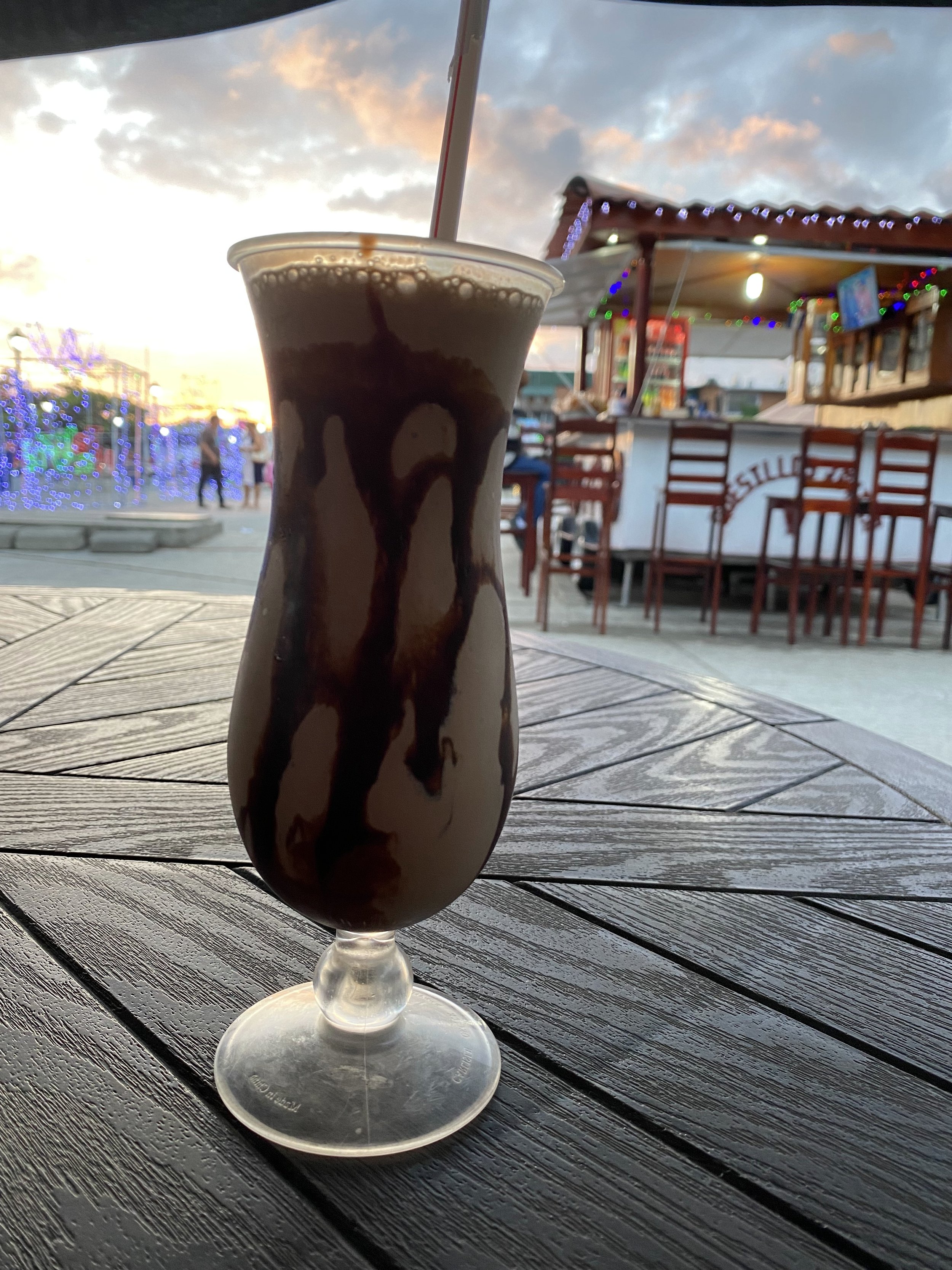 Large chocolate milkshake on an outdoor restaurant table in Rivas, Nicaragua, with sunset skies, Christmas lights, and the town square blurred in the background.